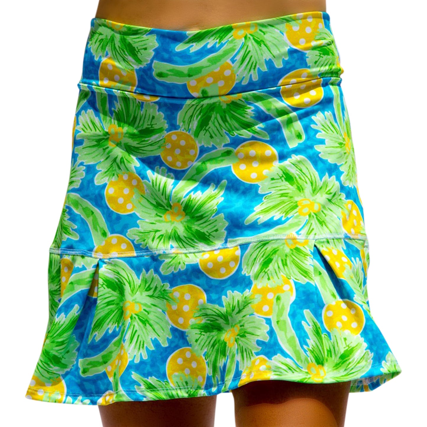 A person wears the Pickleball Bella PB Drop Pleat Skort Palms 1, featuring a blue base with green palm leaves and yellow polka-dotted circles. The flared, ruffled hem shows visible stitching; only the lower torso and upper legs are visible.
