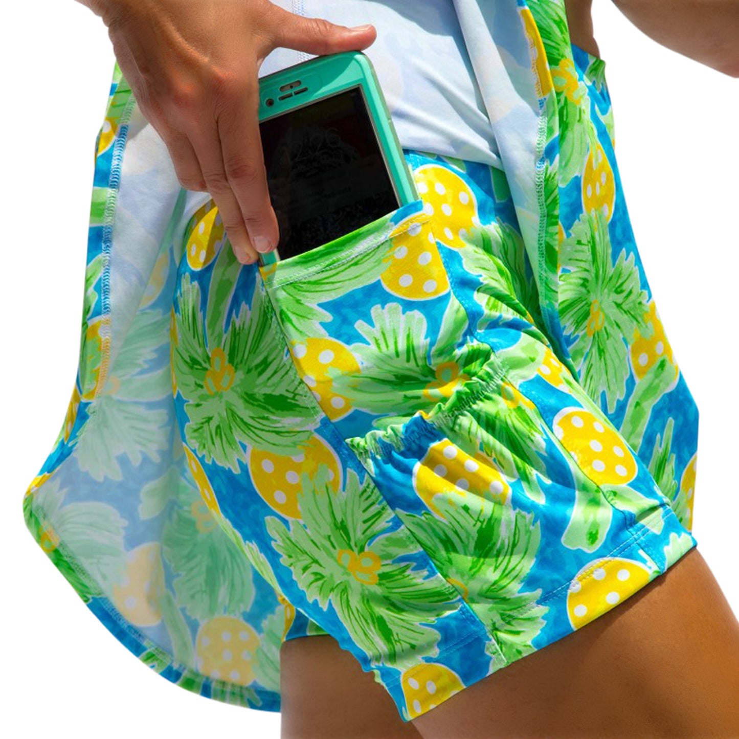 Someone wearing the Pickleball Bella PB Drop Pleat Skort Palms 1, featuring a vibrant coastal print, slips a turquoise smartphone into the skort's pocket as sunlight highlights their leg and the lower edge of a white shirt tucked in.