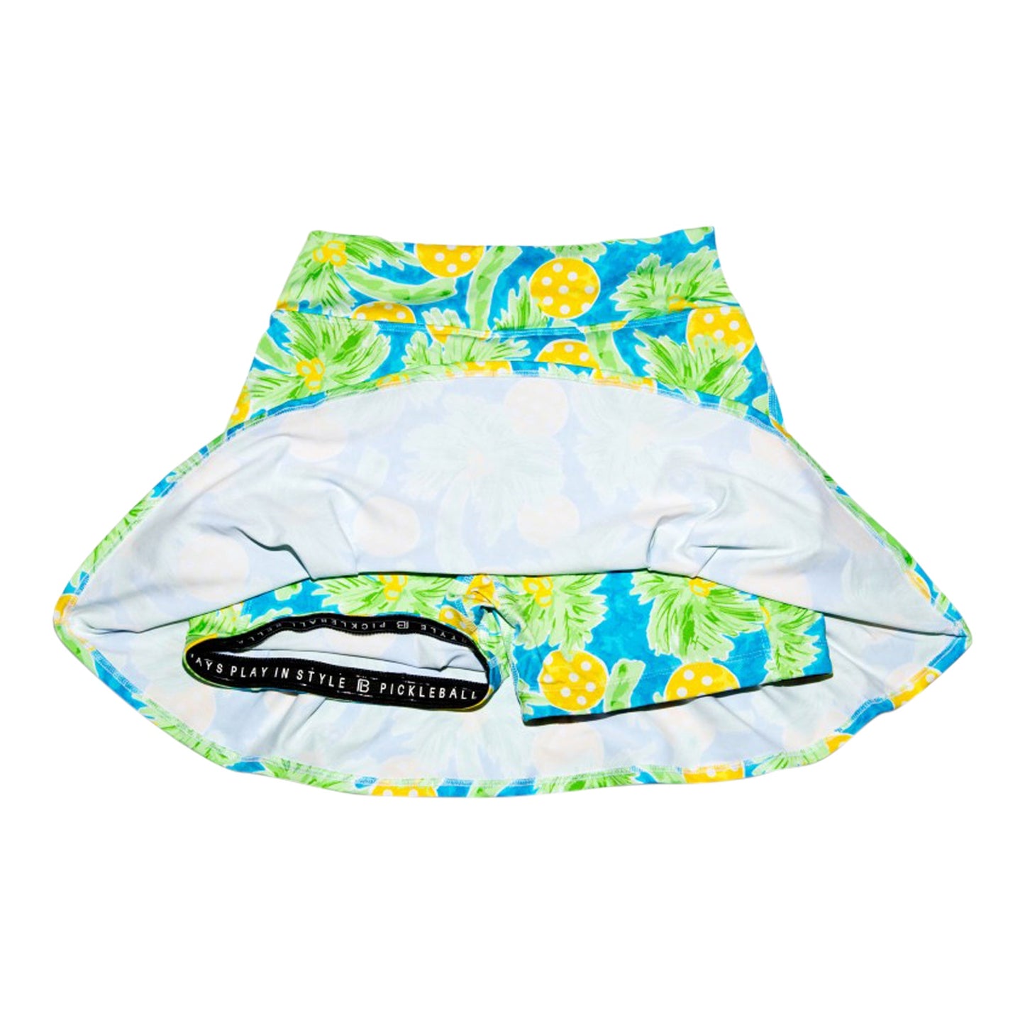 The Pickleball Bella PB Drop Pleat Skort Palms 1 features a vibrant coastal print with yellow, green, and blue pineapples and leaves. Flip it up to reveal fitted compression shorts beneath with a side pocket labeled "PLAY IN STYLE PICKLEBALL.