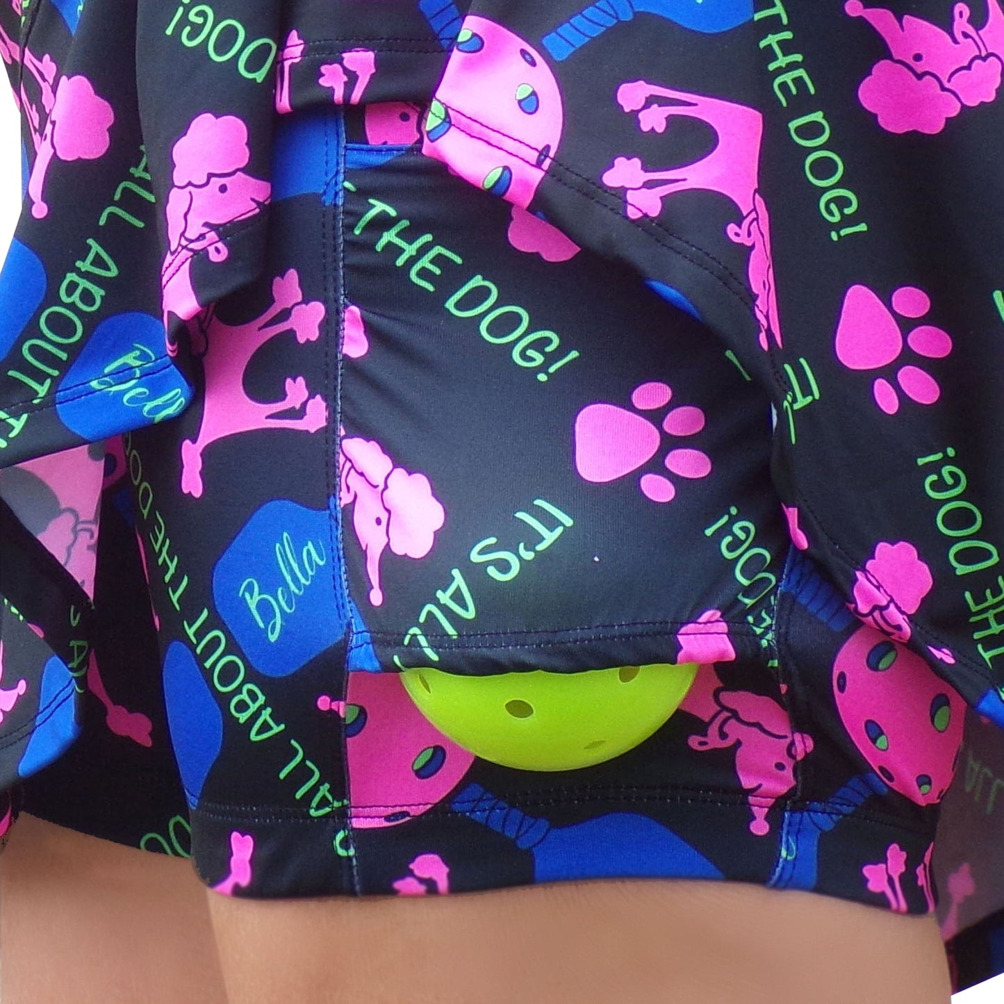 Close-up of the PB Drop Pleat Skort Pickles by Pickleball Bella: black skort featuring colorful designs—pink poodles, blue bones, green tennis balls, pink hearts—and phrases "ITS ALL ABOUT THE DOG!" and "Bella." A green tennis ball peeks from a pocket.