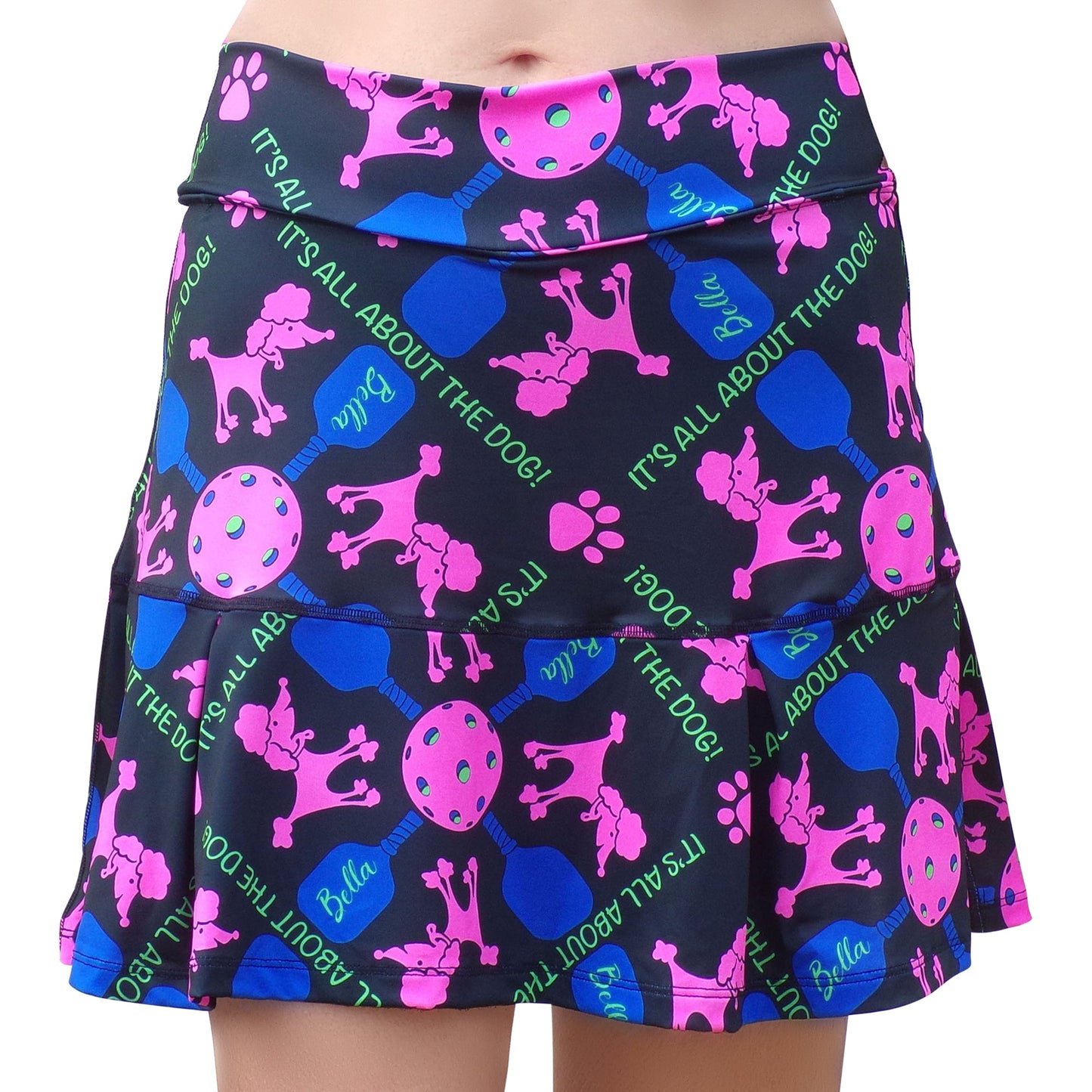 A person wears the Pickleball Bella PB Drop Pleat Skort Pickles, a playful athletic skort with bright pink poodles, blue dog bones, paw prints, tennis balls, green "IT'S ALL ABOUT THE DOG!" text, pleated ruffles, and built-in shorts. Lower torso shown.
