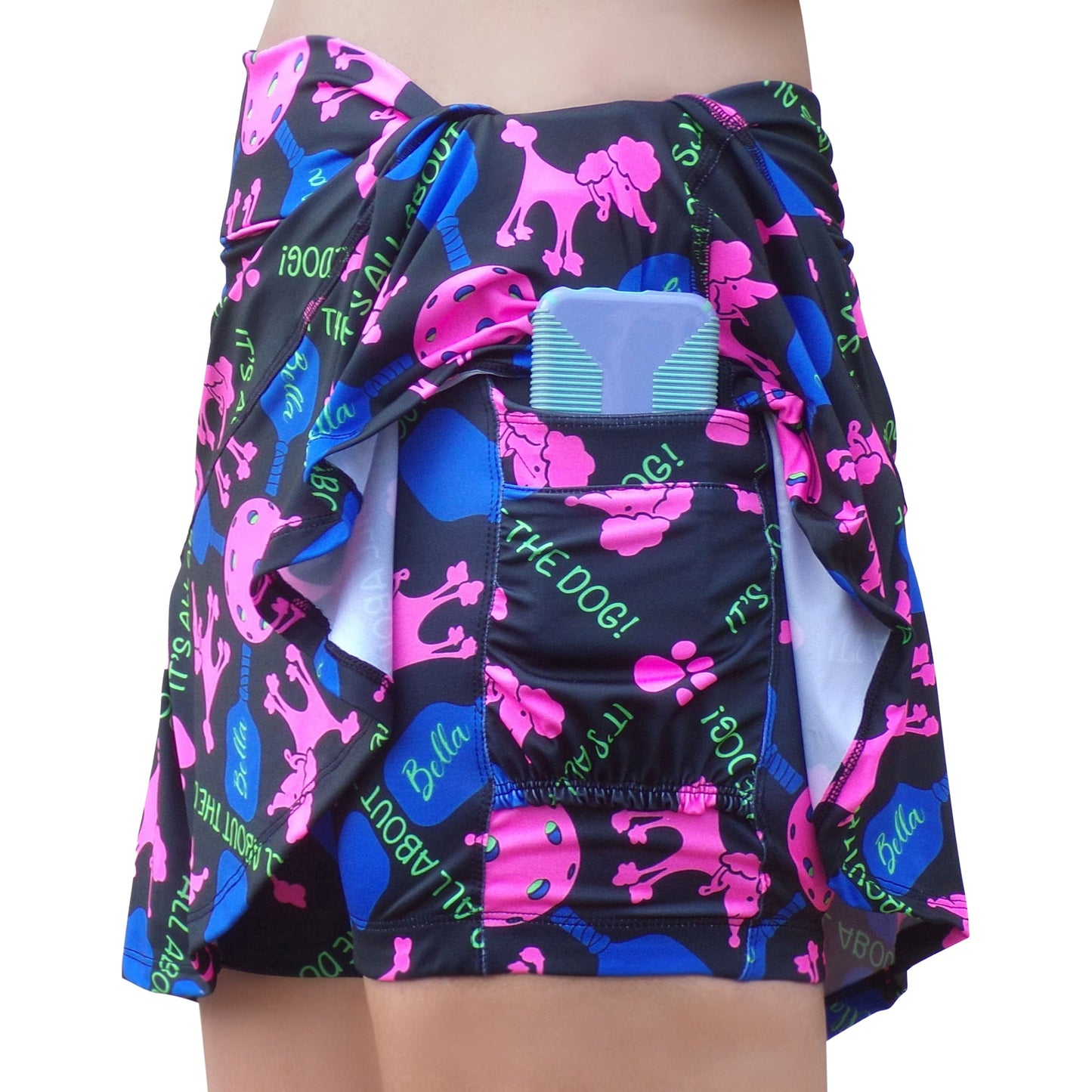 A person wears the PB Drop Pleat Skort Pickles by Pickleball Bella, featuring colorful dog and text patterns. The skort is pulled back to reveal a side pocket holding a rectangular, blue-and-white striped item, possibly a phone.