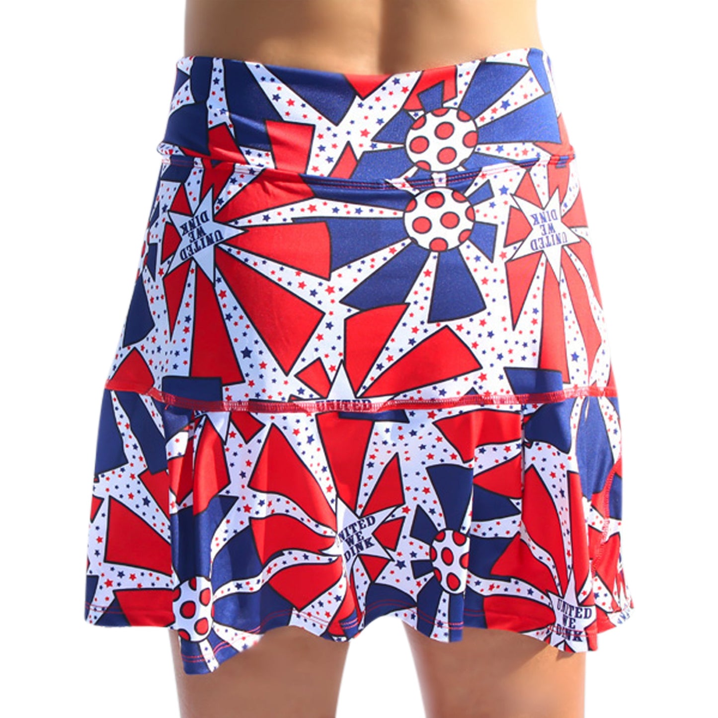 A person wears the Pickleball Bella PB Drop Pleat Skort United We Dink, a patriotic women’s athletic skirt with a red, white, and blue abstract starburst print, white stars, and “United One” text. Photo shows waist to mid-thigh.