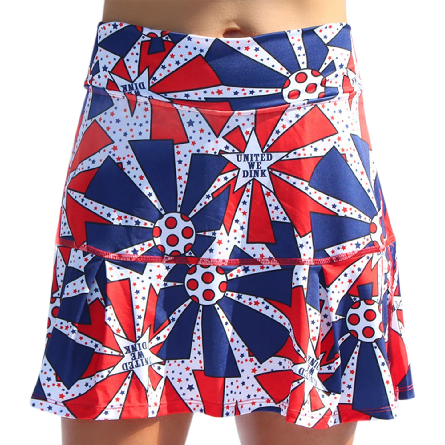 A close-up of someone wearing the Pickleball Bella PB Drop Pleat Skort United We Dink, a women's athletic skirt featuring a bold red, white, and blue starburst pattern with white stars, red circles, and UNITED WE DINK and DINK printed across it.