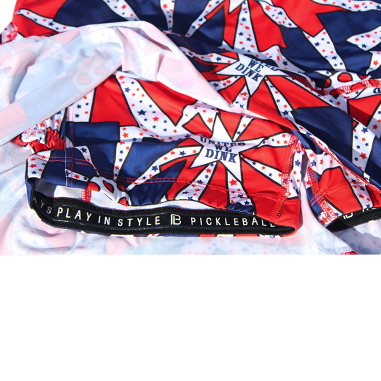A close-up of Pickleball Bella’s PB Drop Pleat Skort United We Dink: red, white, and blue graphics with stars and bold stripes, “PLAYS PLAY IN STYLE PICKLEBALL” waistband, and a “DINK” tag accenting the fabric.