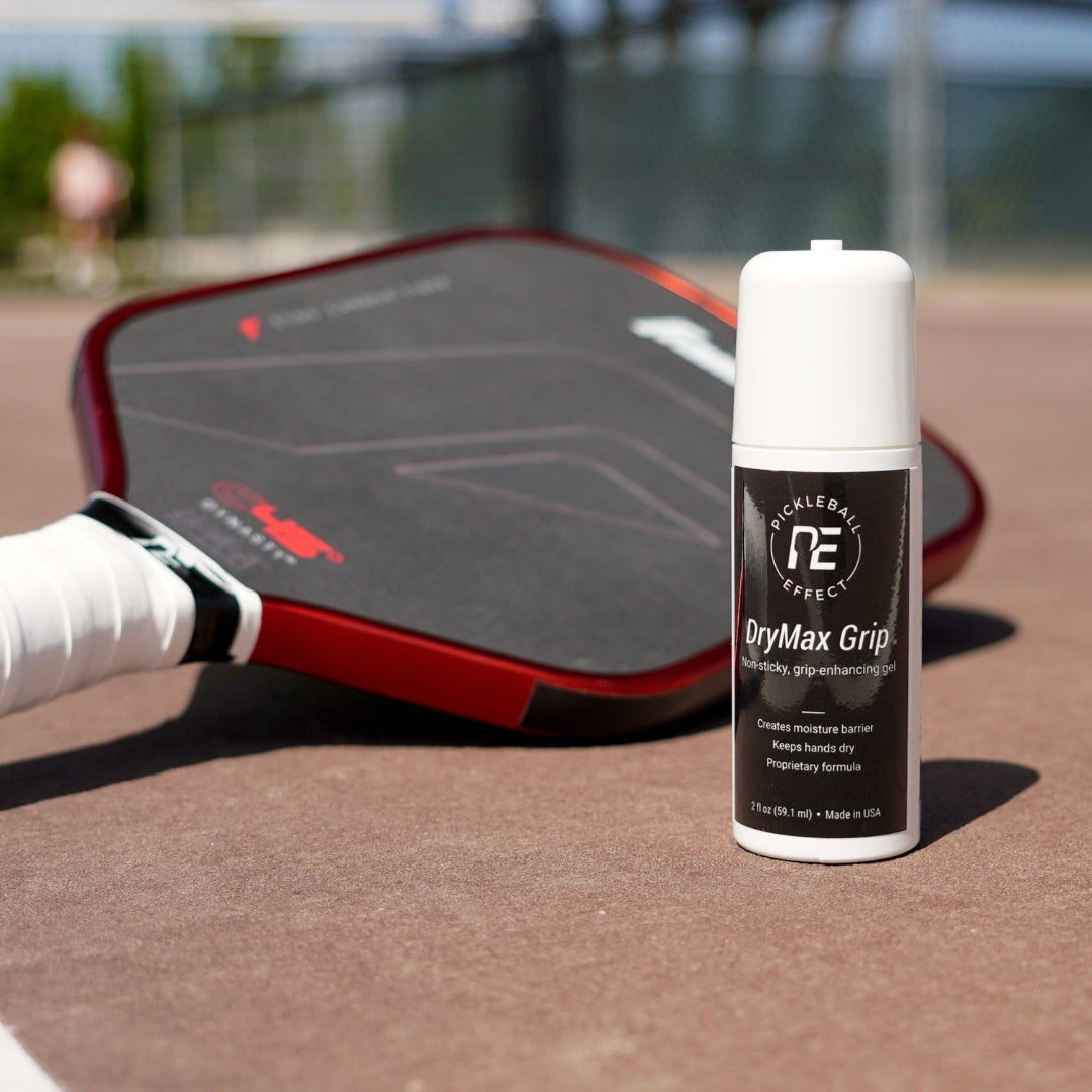 A black and red pickleball paddle sits on an outdoor court, with a small white bottle of DryMax Grip by Pickleball Effect Shop in front—ideal for superior grip and sweat control during sunny games.