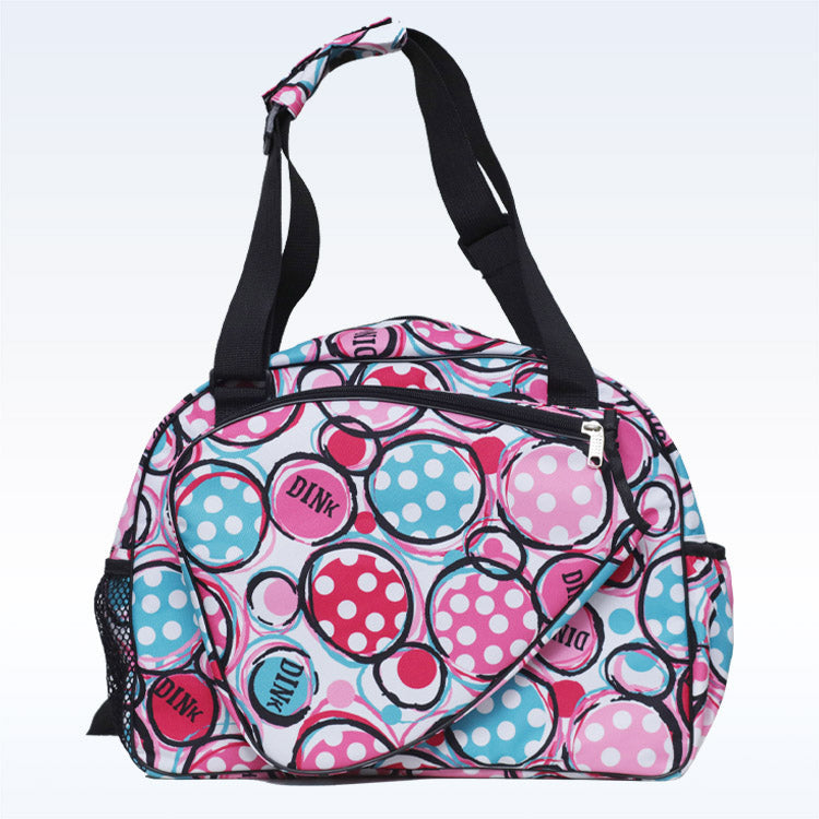 The Pickleball Bella c duffle bag has black straps, a light blue background, overlapping blue, pink, and red circles with white polka dots—some labeled DINK—and features zippered paddle and front pockets plus a side mesh pocket.