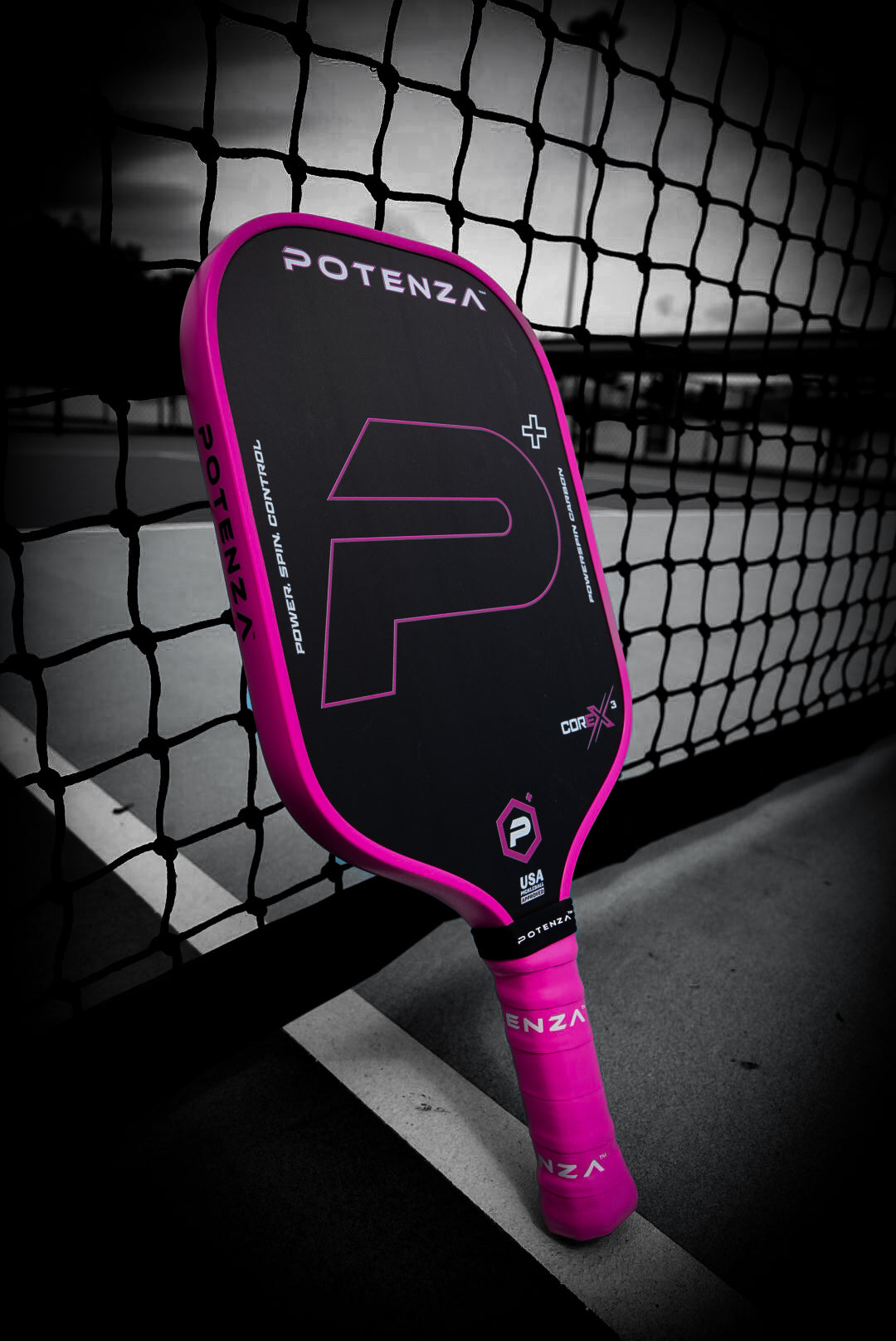 A P+ PowerSpin Carbon COREx3 paddle (Neon Pink, Elongated Face) from Potenza Pickleball leans against a net on an outdoor court. The desaturated background highlights the paddle’s vivid pink accents, with court lines and net in view.