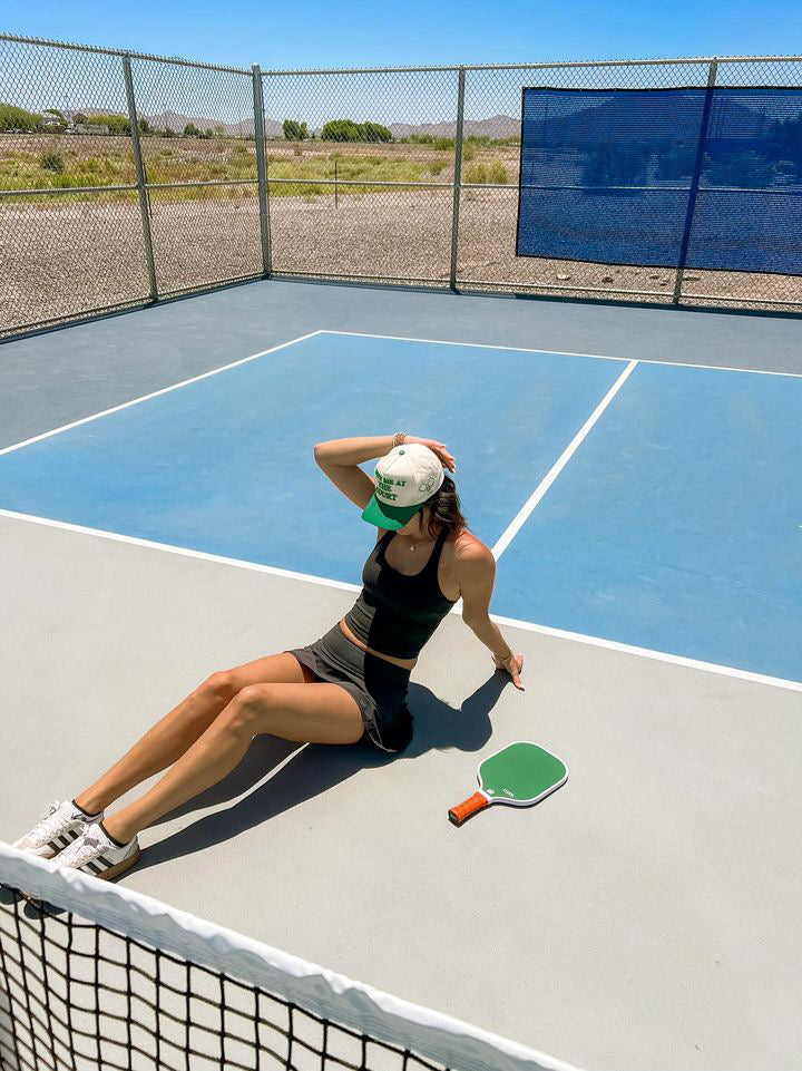 A woman in a black athletic outfit and white sneakers sits on a pickleball court next to a green paddle, resting one hand on the ground and the other on her LUXE Pickleball Meet Me At The Court Hat under the sunny blue sky, framed by a chain-link fence.