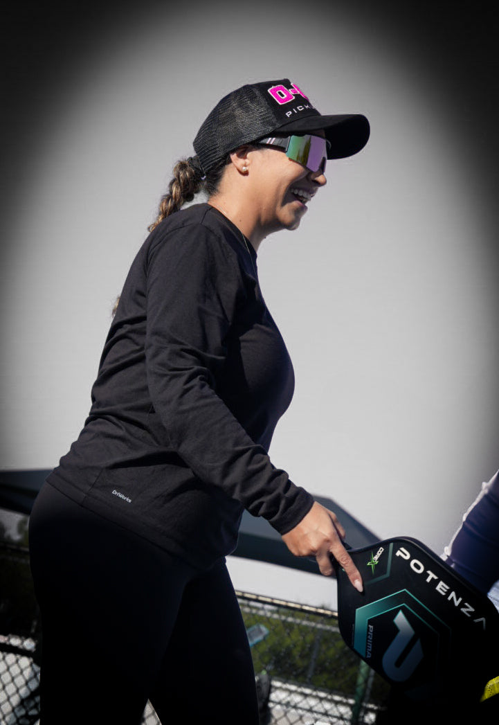 A woman in black athletic wear and sunglasses smiles outdoors, wearing the Potenza Pickleball "0-0-2" Collection Hat (0-0-START) with chenille embroidery, and holding a Potenza pickleball paddle. The sunny background is blurred with a fence.