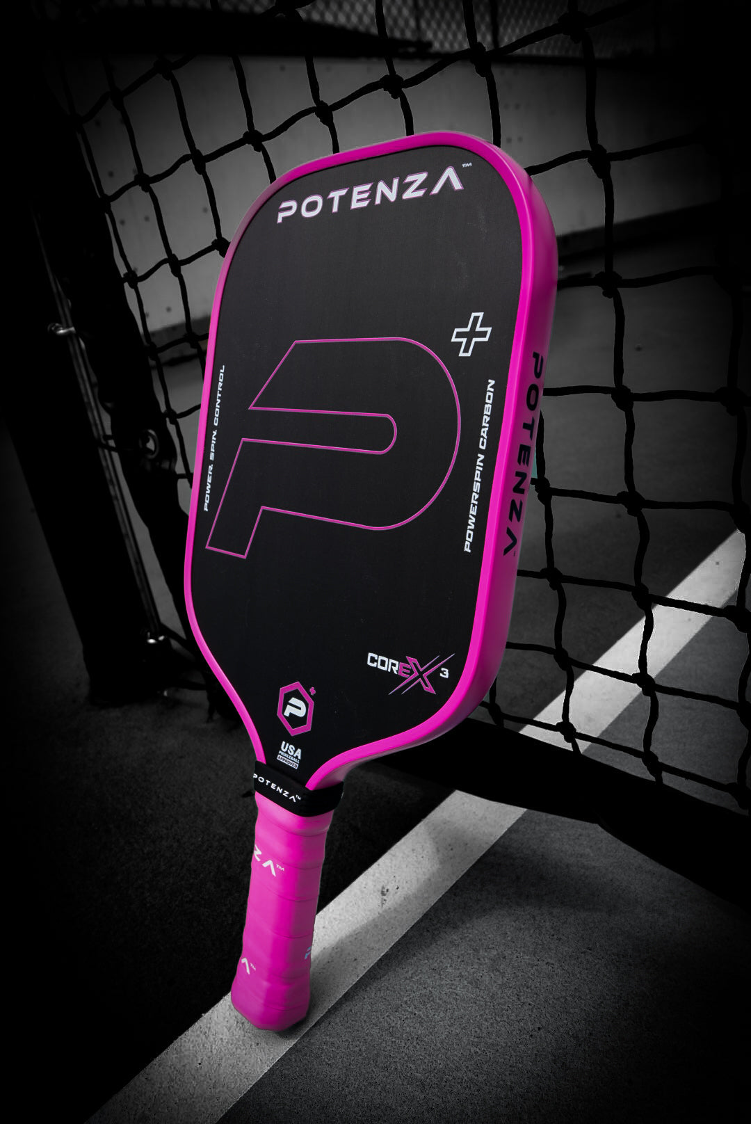 A Potenza Pickleball P+ PowerSpin Carbon COREx3 (Neon Pink, Elongated Face) paddle with vibrant pink edges and grip, and a bold pink "P" on its carbon fiber face, leans against a black net on an outdoor court.