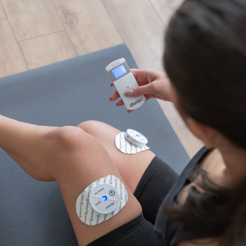 A person sits on a yoga mat indoors, using the iReliev Therapeutic Wearable System with two electrode pads on their left knee and thigh, holding the handheld device for pain relief or muscle stimulation. A wooden floor is visible in the scene.