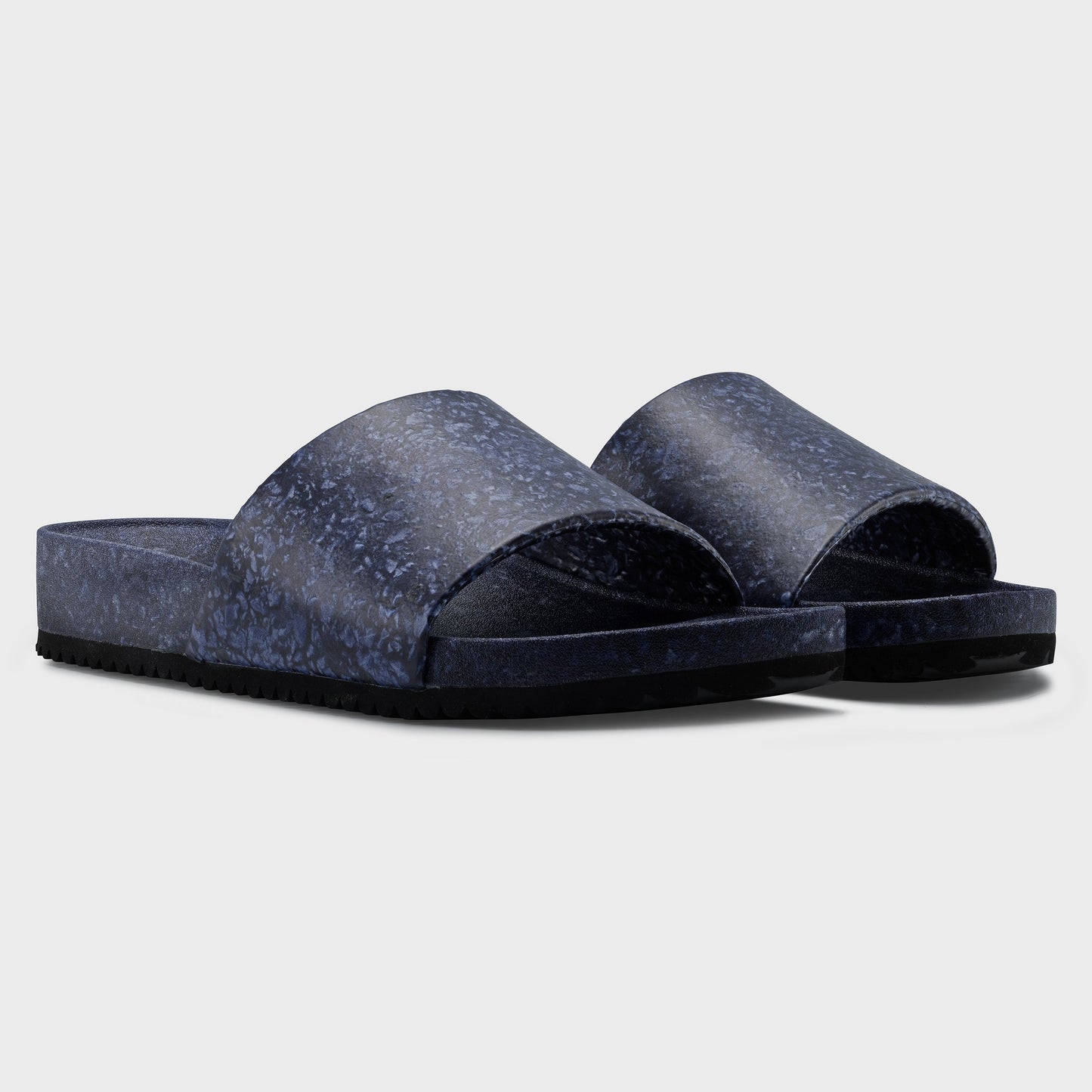 The Blumaka Fleks® East Beach Slide features navy blue marbled textured wide straps and slightly raised soles for alignment support. Shown on a light gray background, these sandals combine style with comfort.