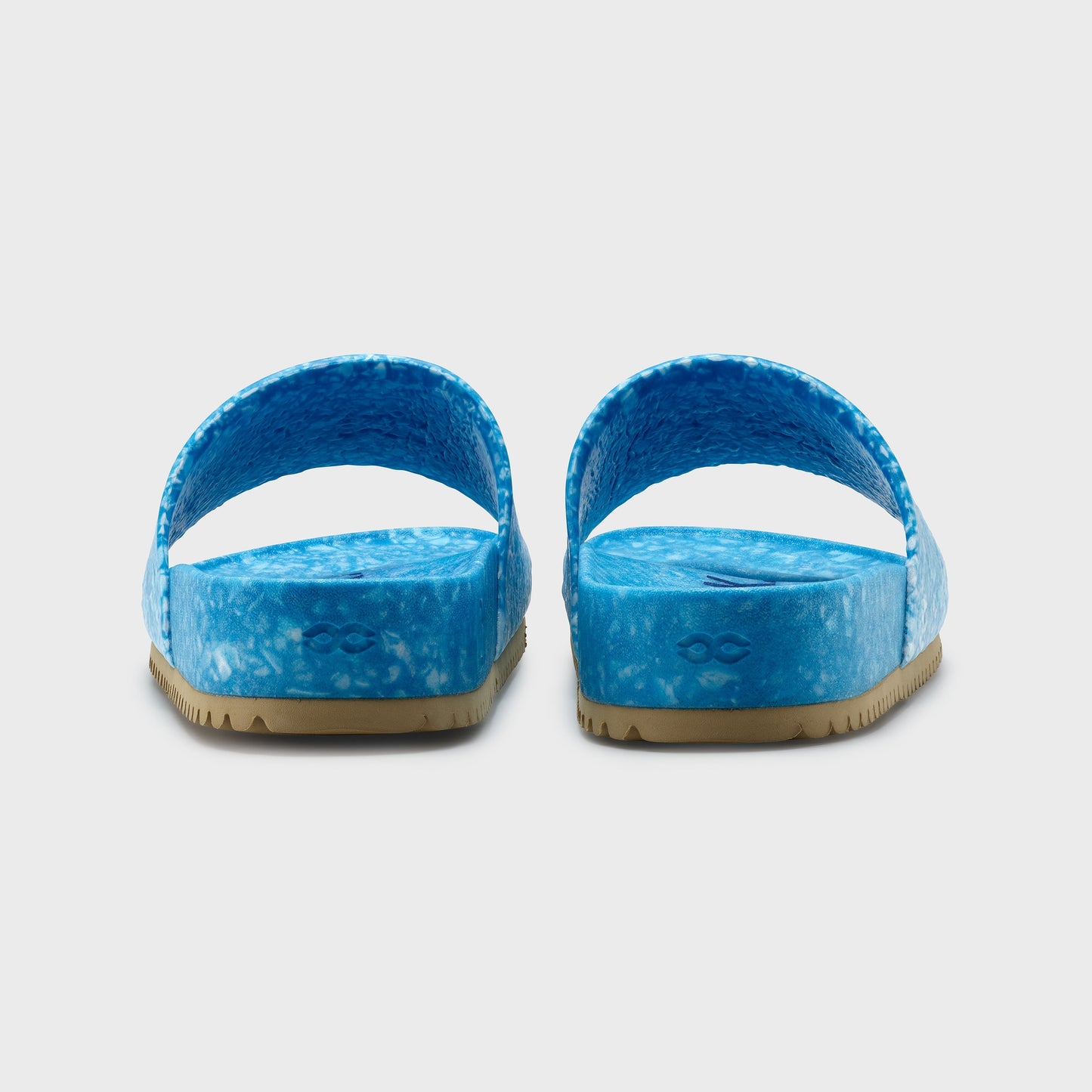 The Blumaka Fleks® East Beach Slide, shown from the back against a white background, features bright blue marbled slip-on design, wide straps for support, flat tan soles with wavy tread, and is made to promote proper alignment.