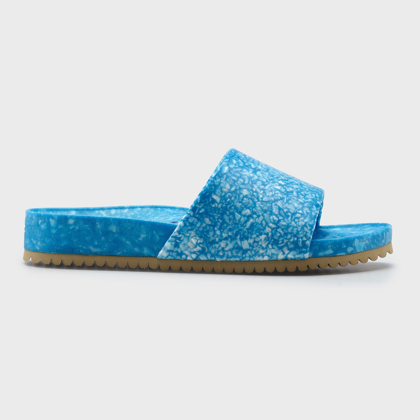 The Blumaka Fleks® East Beach Slide is a blue, slip-on sandal with a marbled finish, wide strap, and thick flat sole with light brown jagged tread—ideal for alignment support and post-athlete recovery. Shown in profile on white.