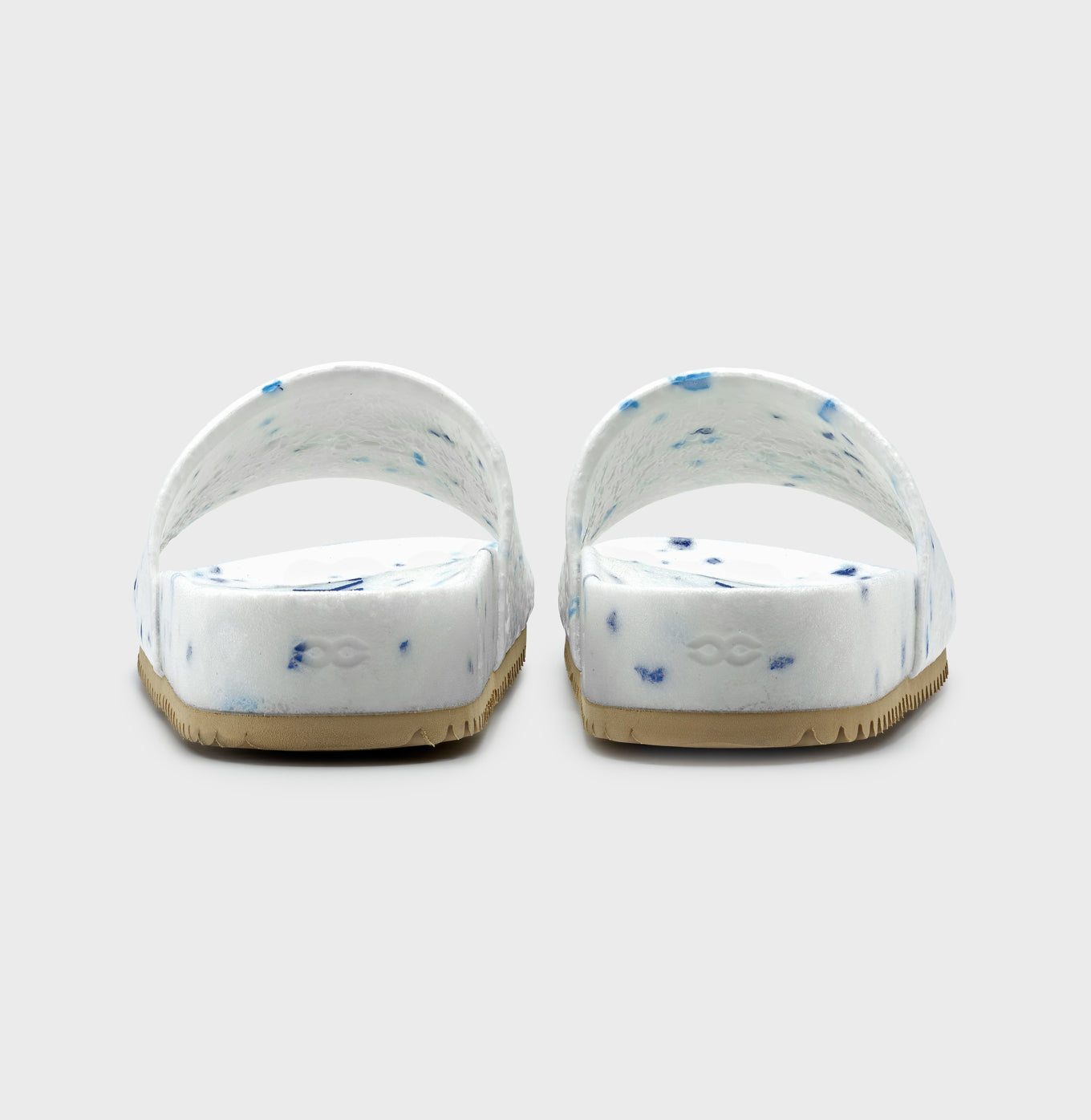 The Blumaka Fleks® East Beach Slide sandals, seen from the back, feature a white design with blue speckles, wide straps, and tan textured soles for alignment support—all set against a plain light gray background.