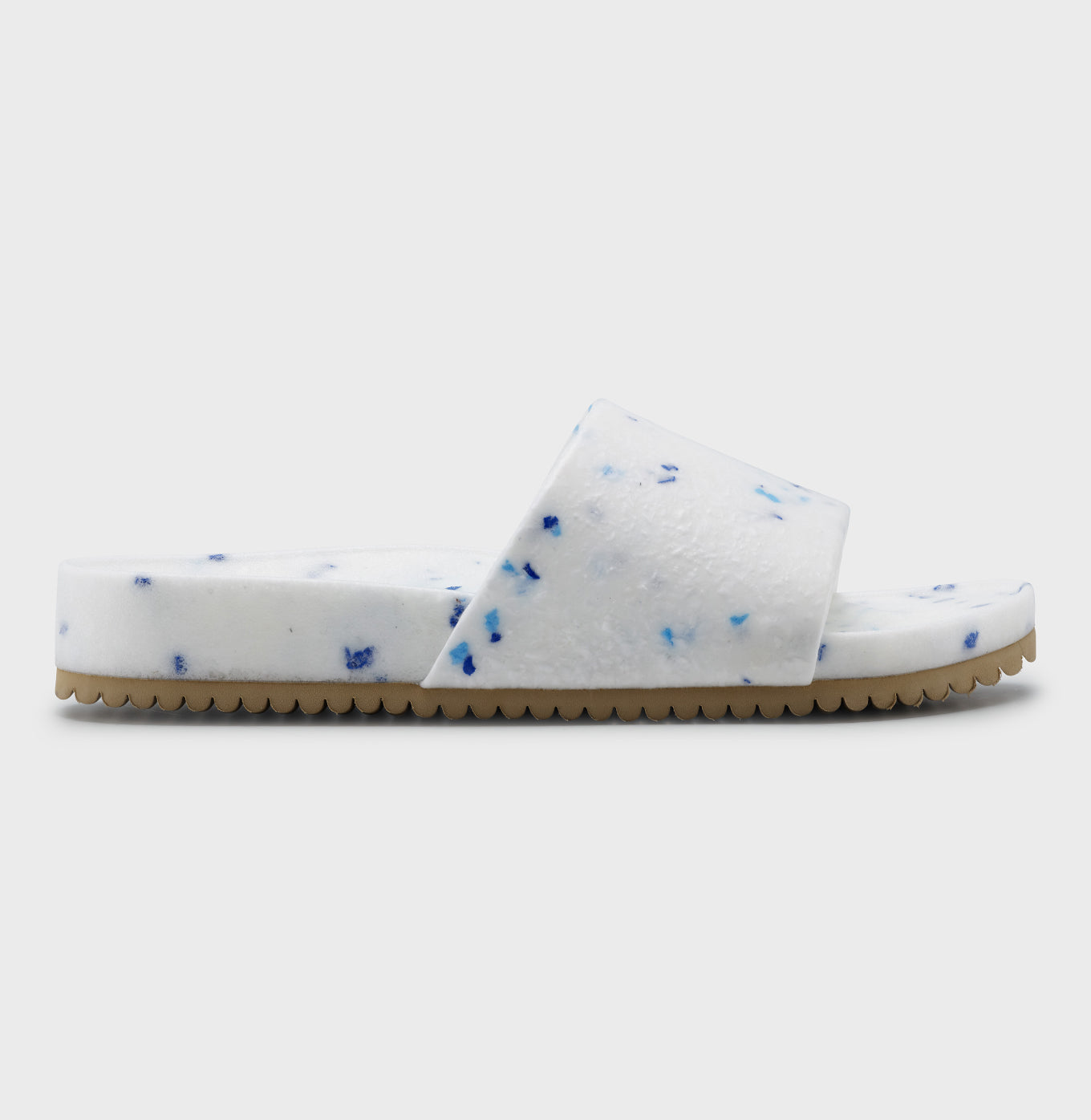 The Blumaka Fleks® East Beach Slide features a fuzzy white upper with blue speckles, thick strap, plush comfort, and flat tan ridged outsole for support and alignment—ideal for athlete recovery. Shown on a plain light gray background.