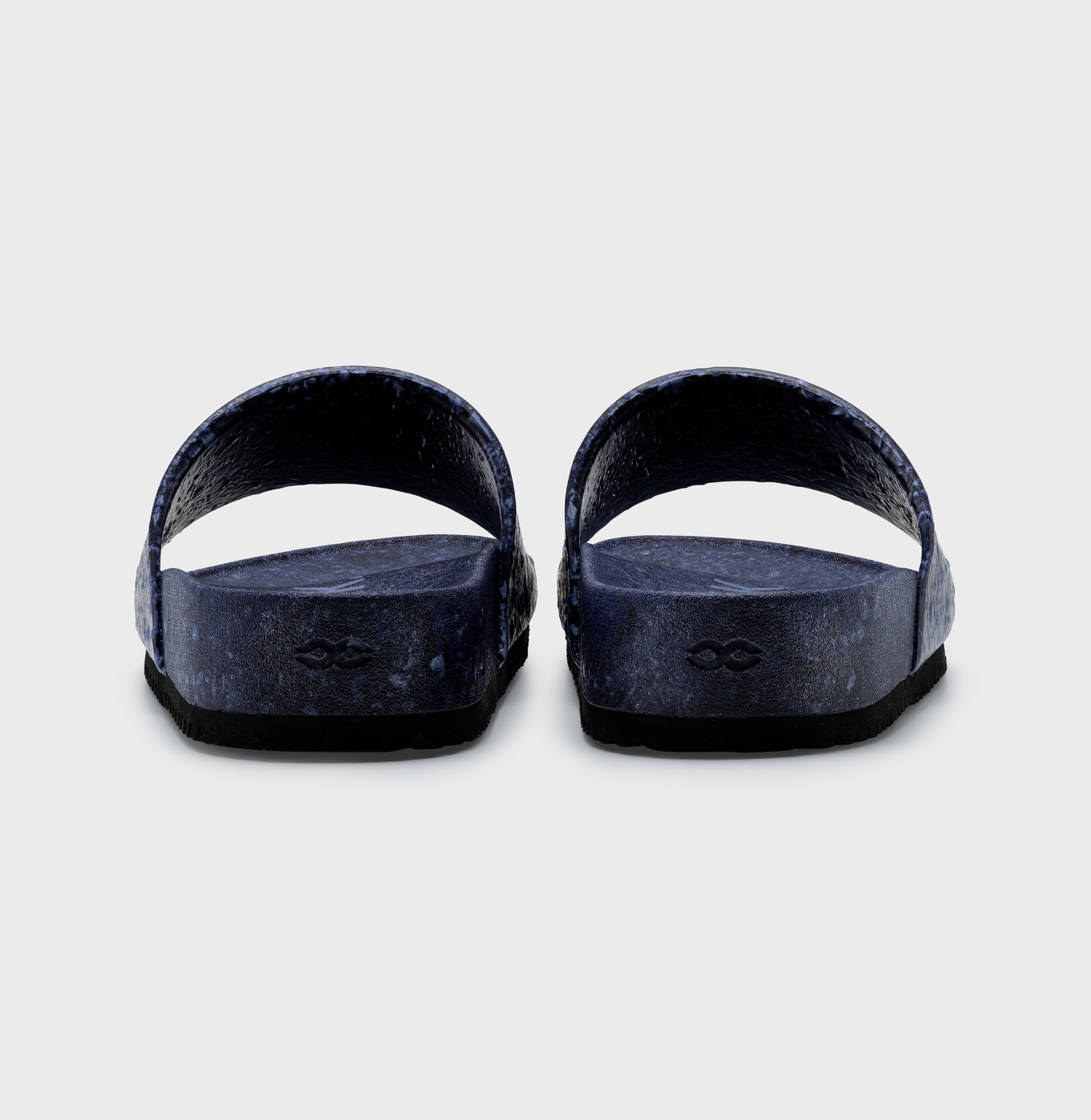 The Blumaka Fleks® East Beach Slide sandals, in dark blue with textured single wide straps and contoured footbeds, are shown from the back on a light gray background, each heel featuring a small logo imprint for subtle branding.