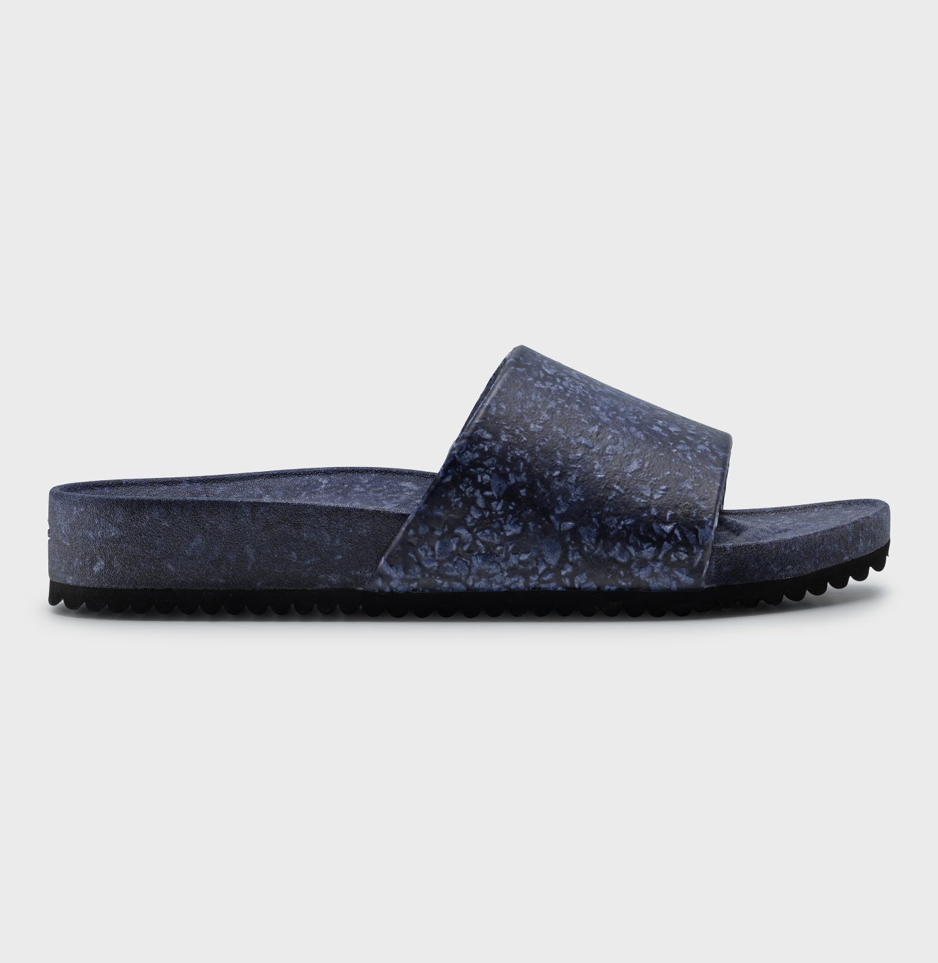 The Blumaka Fleks® East Beach Slide is a dark blue slip-on sandal with a textured upper strap and sole, featuring a flat, slightly raised heel for athlete recovery and alignment. Shown side view against a plain white background.