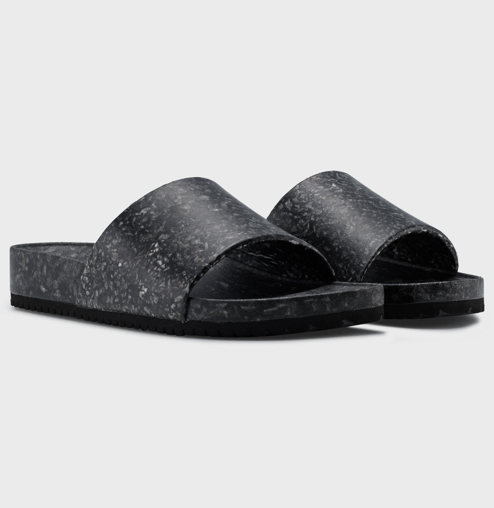 The Blumaka Fleks® East Beach Slide, featuring a wide marbled strap and thick flat sole for alignment support, is displayed on a light gray background at an angle highlighting both the side and front views.