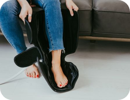 A person in blue jeans sits on a sofa, placing their bare, red-painted feet into the iReliev Compression Therapy Bundle. The black device is plugged in with a white cord, resting on a light-colored floor to help improve circulation.