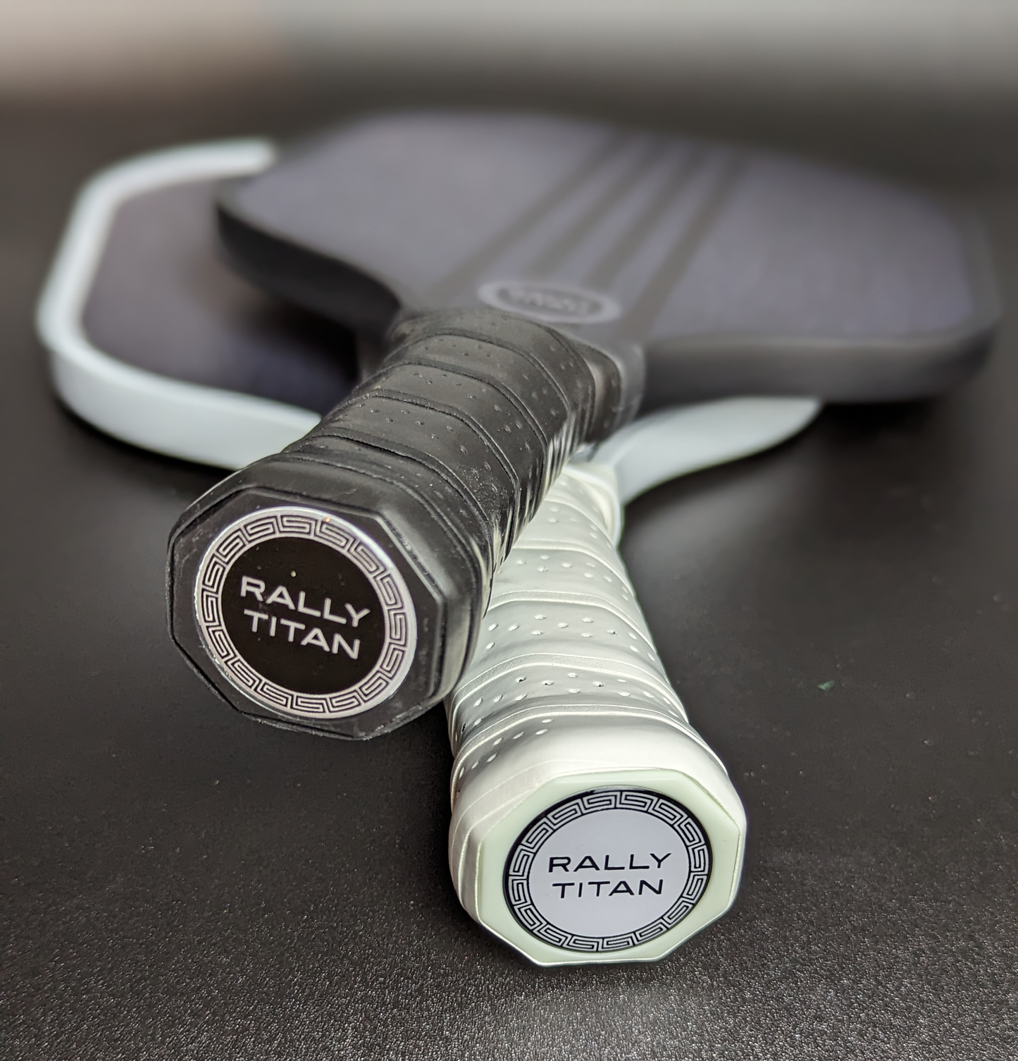 Close-up of two Rally Titan Pickleball Aspro 10MM paddles with T300 textured carbon fiber surface, featuring black and white grips and bold brand lettering on the end caps, lying on a dark surface.