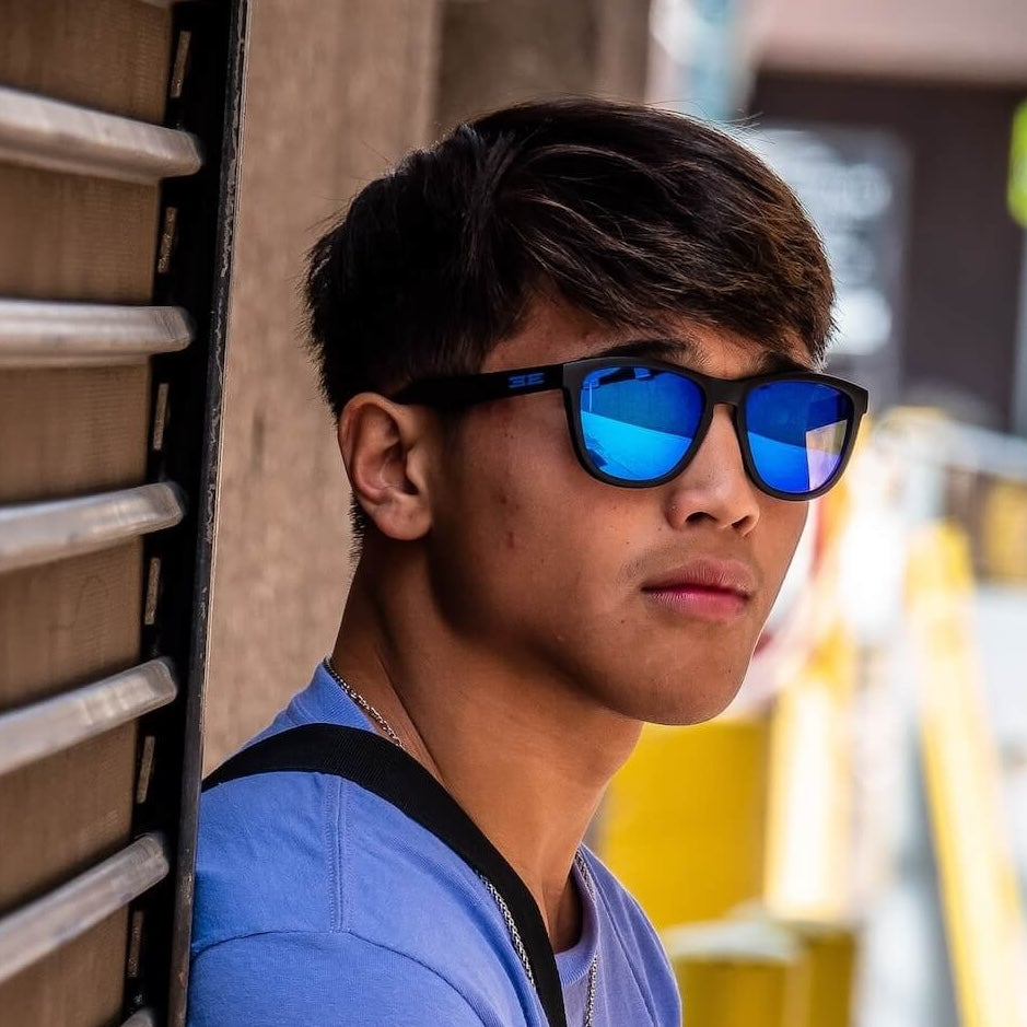 A young man with short dark hair wears reflective blue VIBE sunglasses by Epoch Eyewear and a light blue shirt. He leans against a metal shutter outdoors, looking away with a neutral expression as bright yellow barriers blur in the background.