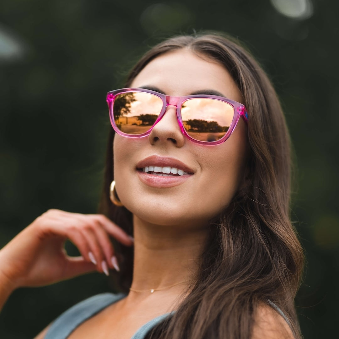 A woman with long brown hair smiles at the camera, wearing Epoch Eyewear VIBE pink polarized sunglasses with mirrored lenses. She has gold hoop earrings and manicured nails, standing against a blurred green outdoor background.