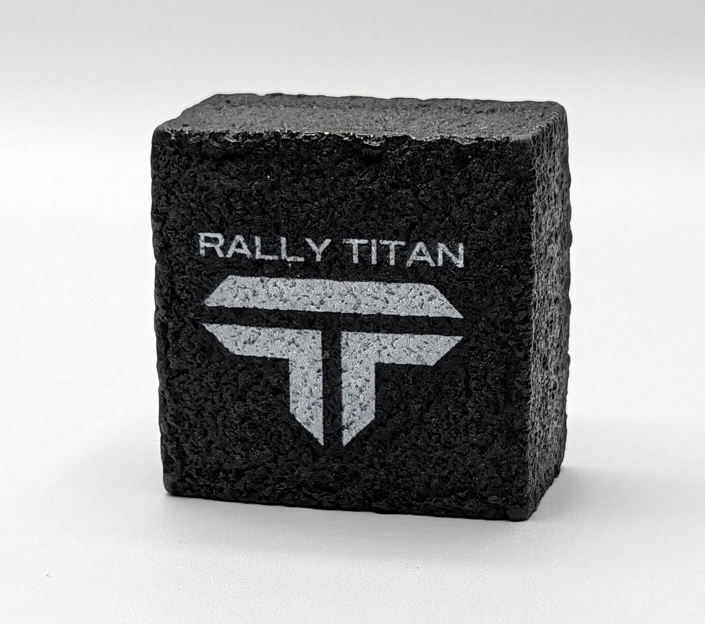 A textured black rectangular block, the Rally Titan Pickleball Paddle Eraser, stands upright on a white surface. Its front features "RALLY TITAN" and a bold "T" logo, with its rough surface designed to clean pickleball paddles effectively.