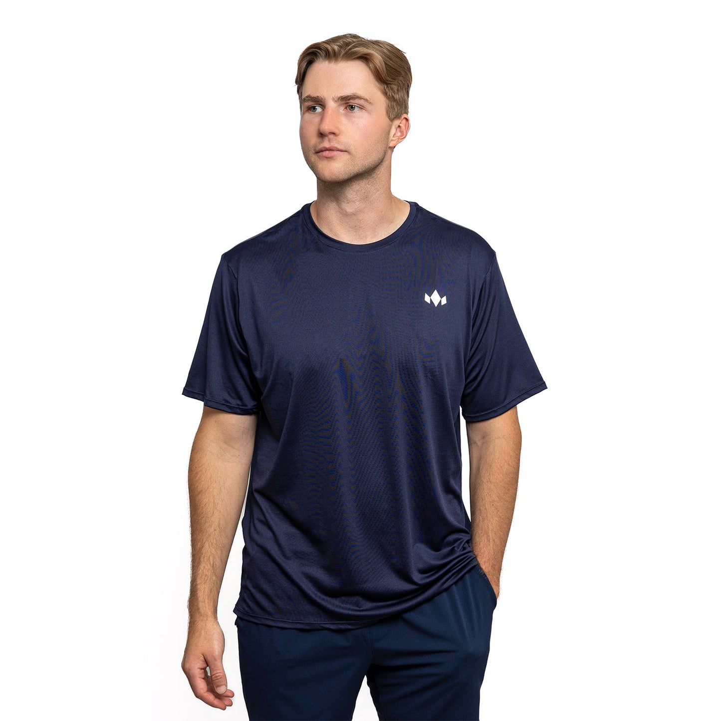 A man with light brown hair wears the Diadem Sports - Men's Essential Everyday Top, a navy blue short-sleeve shirt by Diadem Sports, featuring an athletic fit and performance-driven comfort, standing with one hand in his pocket against a white background.
