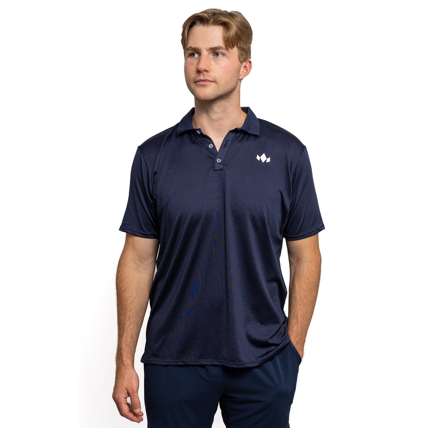A man with light brown hair stands against a white backdrop, wearing a navy blue Diadem Sports - Men's Essential Everyday Top, an athletic-fit short-sleeve polo with a small white geometric logo on the chest and one hand in his pocket.