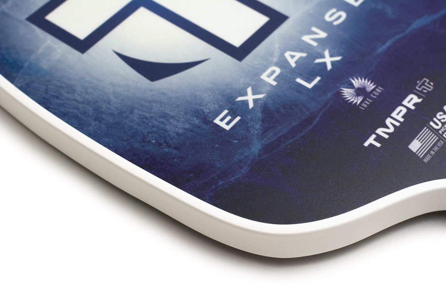 Close-up of the TMPR Sports Expanse LX | Indigo Ice paddle’s corner, highlighting its “EXPANSE LX,” “TMPR,” and “USA Pickleball” logos. The wide-body paddle displays its textured surface, curved white edge, and built-in vibration dampening.