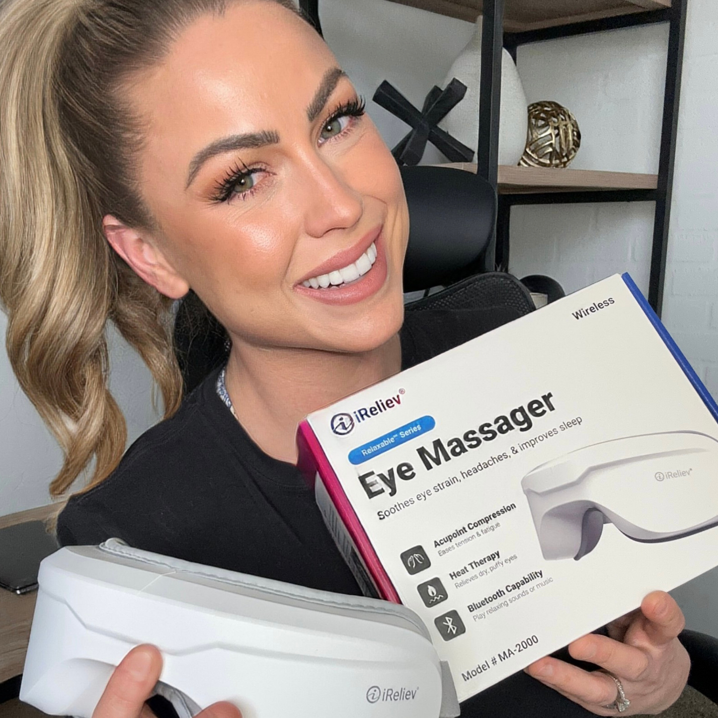 A smiling woman with long hair holds the iReliev Relaxable™ Eye Massager with Heat in one hand and its box in the other. The box highlights features like compression therapy, heat therapy, and Bluetooth capability.