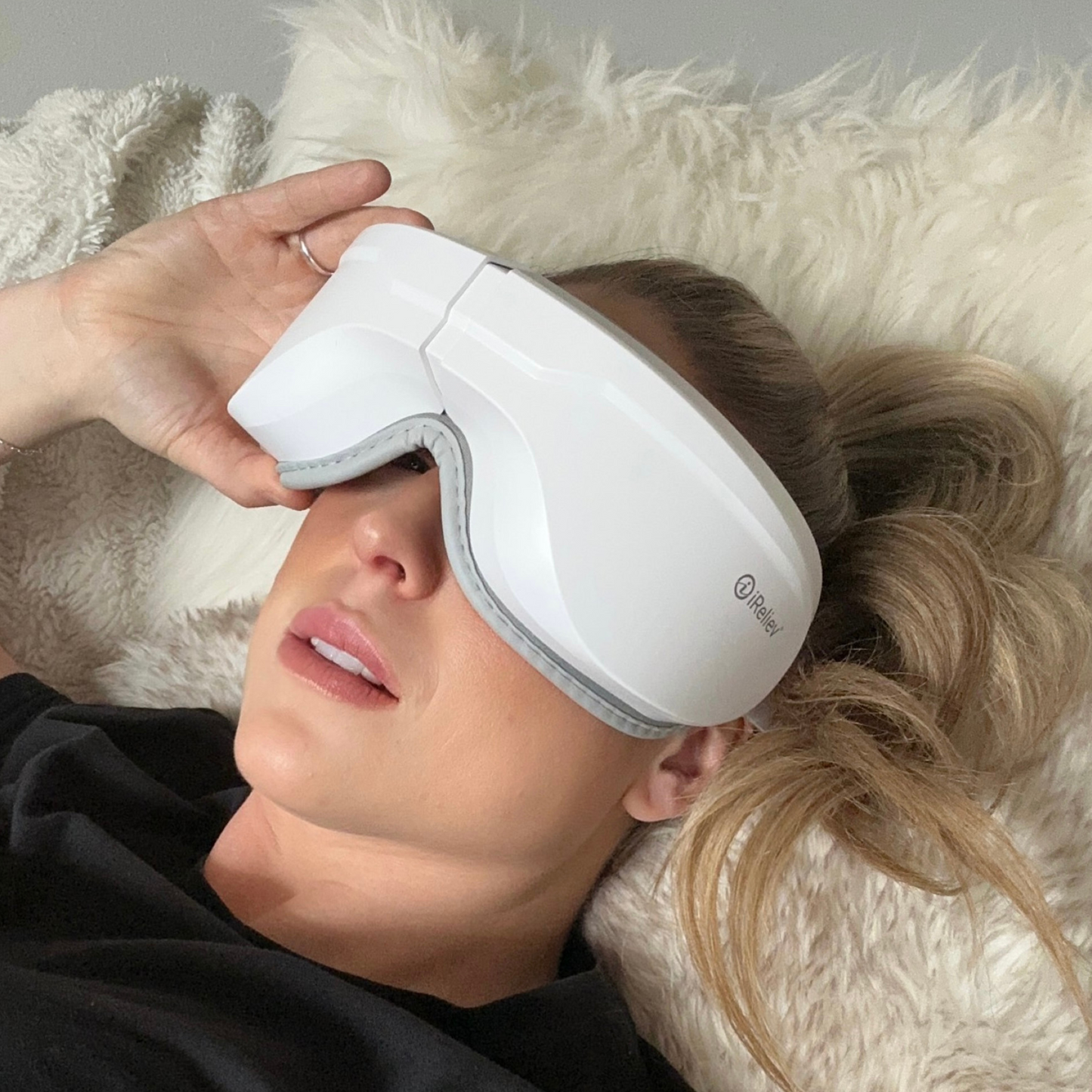 A woman in a black shirt lies on a fuzzy cream blanket, wearing the iReliev Relaxable™ Eye Massager with Heat over her eyes and resting her hand on it for soothing compression therapy.