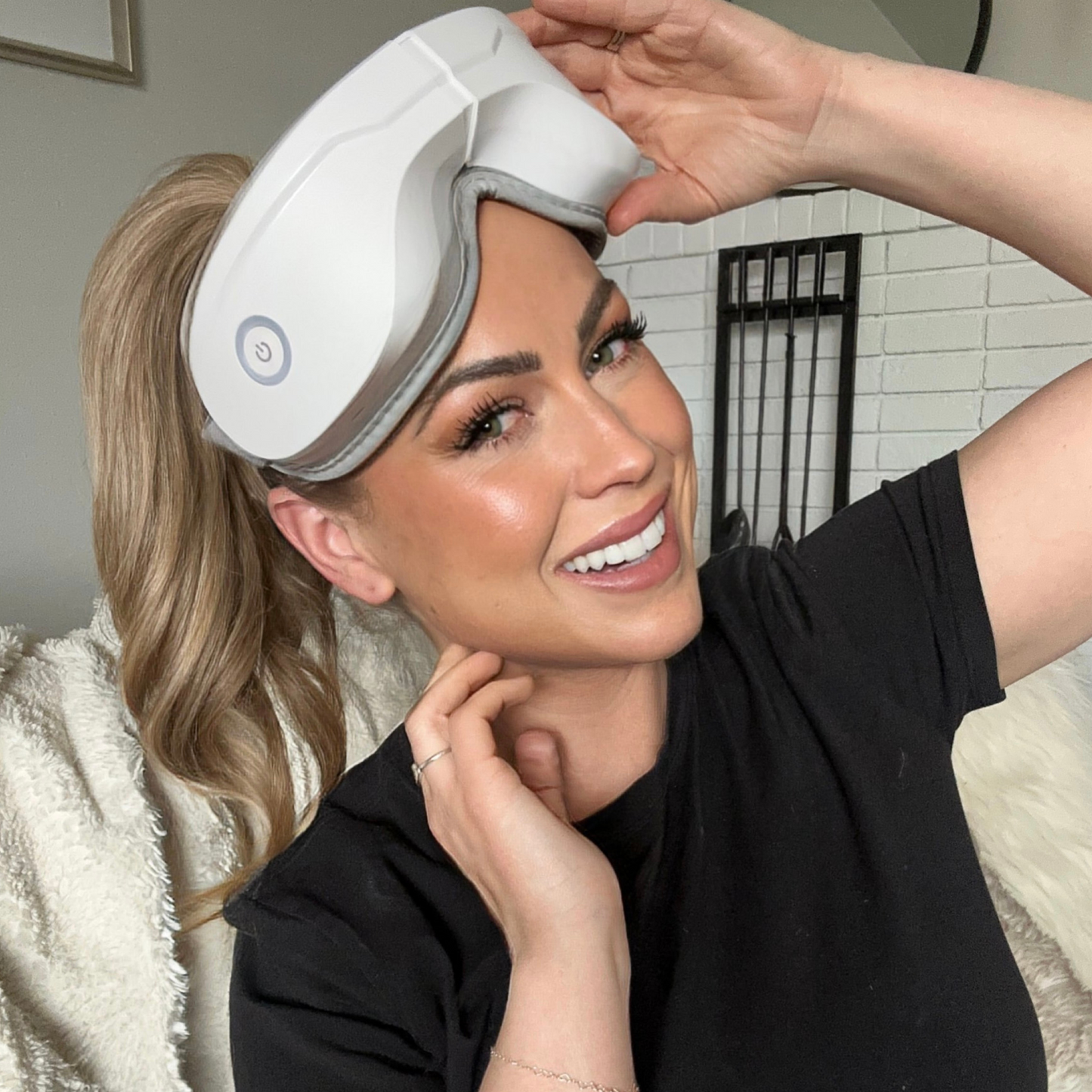 A smiling woman with wavy blonde hair, wearing a black t-shirt, adjusts the iReliev Relaxable™ Eye Massager with Heat on her forehead while sitting on a couch with a cream blanket against a white brick wall.