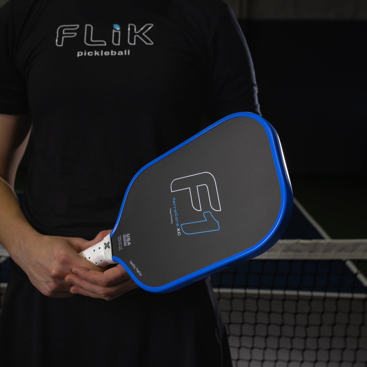 Wearing a black “FLiK pickleball” shirt, a person holds the USAP-approved body helix FLiK F1 Elongated Pickleball Paddle with a white grip, featuring black and blue colors, in front of an indoor net, with focus on the paddle.