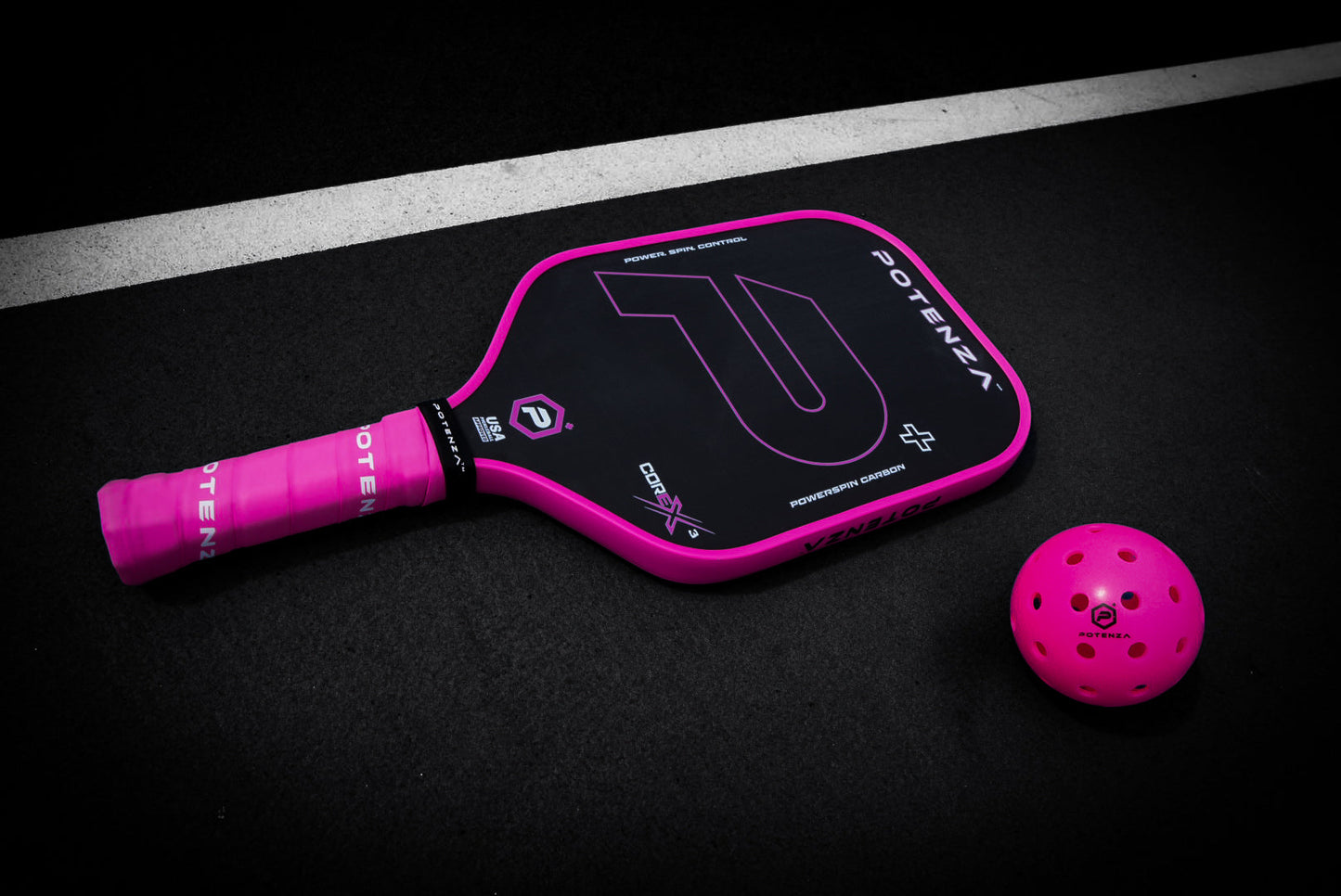 A Potenza Pickleball P+ PowerSpin Carbon COREx3 paddle in neon pink and black rests on a black court near a white line, with a matching pink ball—both designed for optimal spin on textured surfaces.