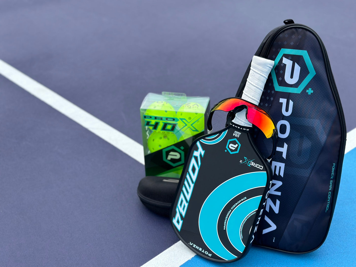 A Potenza Pickleball paddle with a black and teal design lies on a blue court next to its POTENZA Paddle Travel Case (fits 2 paddles), neon green pickleballs, and red-lensed sunglasses near white court lines.