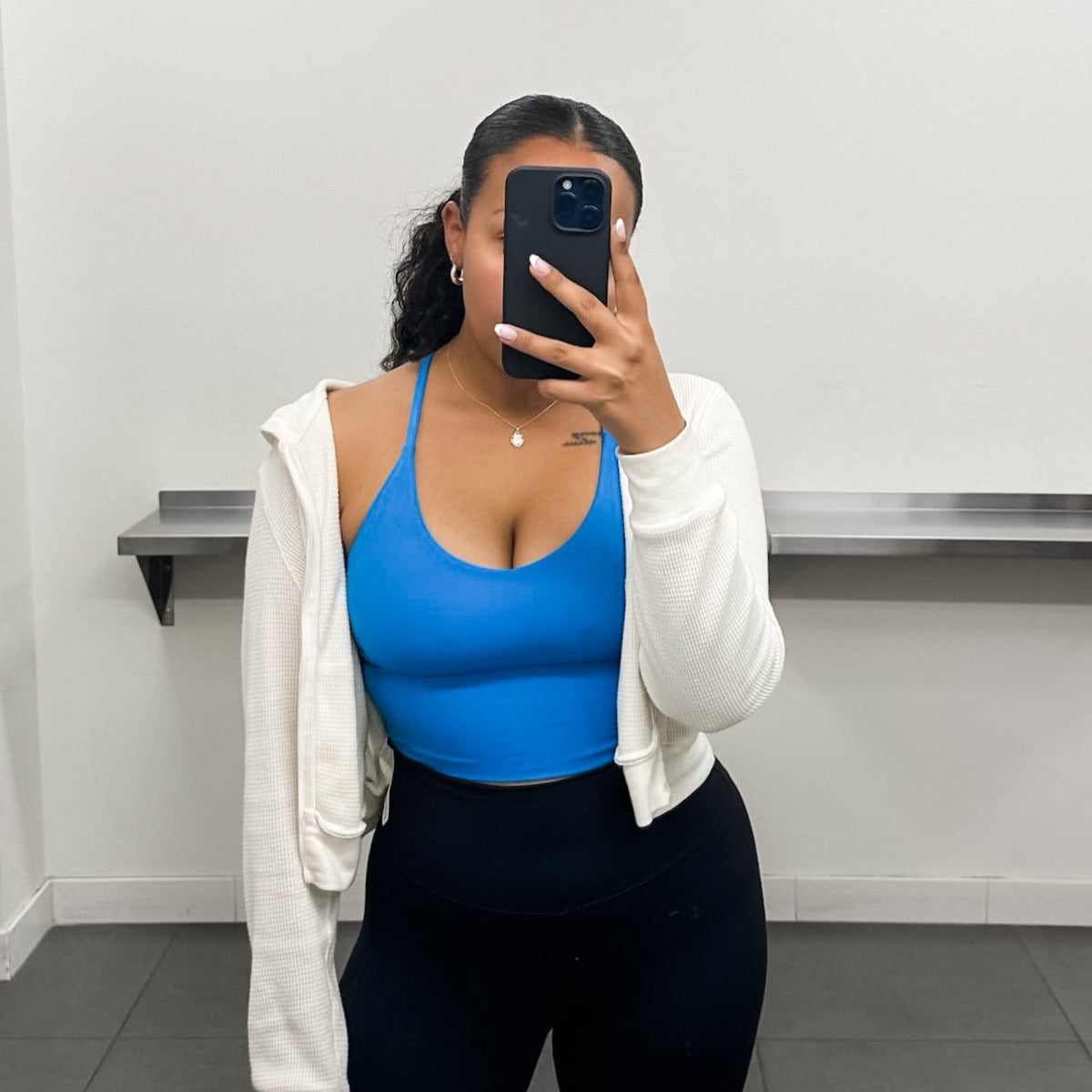 A woman takes a mirror selfie in a minimal room, wearing the DYM Athletics Flex & Flow Racerback Tank in blue, paired with black leggings and a white open cardigan. Her hair is tied back, face hidden by her phone, with a metallic shelf behind her.