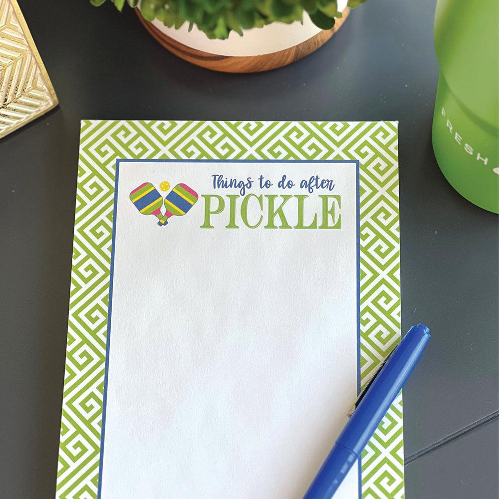 The Fresh Pickle Pickleball Notepad features “Things to do after PICKLE” with crossed paddles and a ball, plus a green geometric border—ideal for pickleball fans or as a unique gift. Blue pen, drink bottle, and plant not included.
