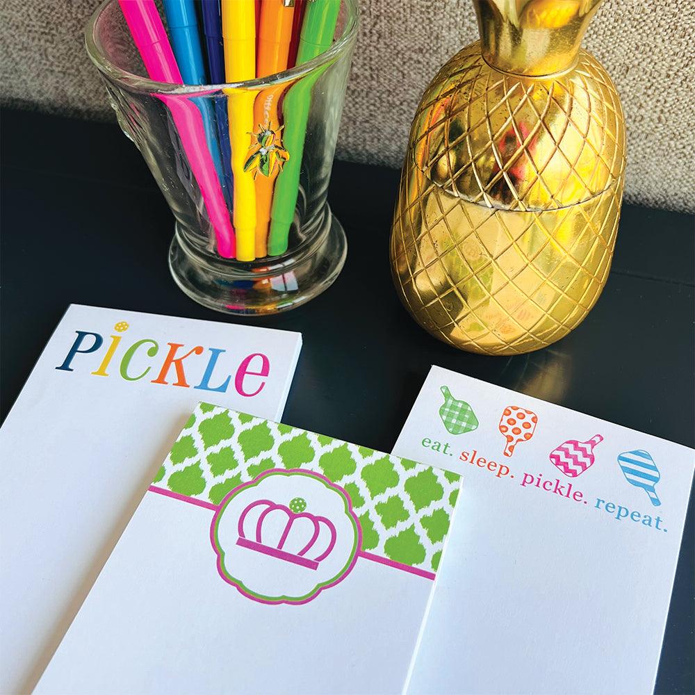 A desk displays a glass of pens, a gold pineapple container, and three Fresh Pickle Pickleball List Notepads featuring fun phrases like “eat. sleep. pickle. repeat.”—perfect gifts for any pickleball lover.