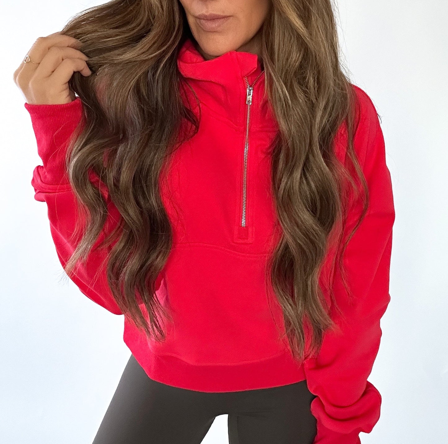 A woman with long, wavy brown hair models the DYM Athletics Cozy Up Funnel Neck 1/2 Zip Hoodie in bright red with dark leggings. Against a plain white background, she touches her hair, showing only her lips and chin.
