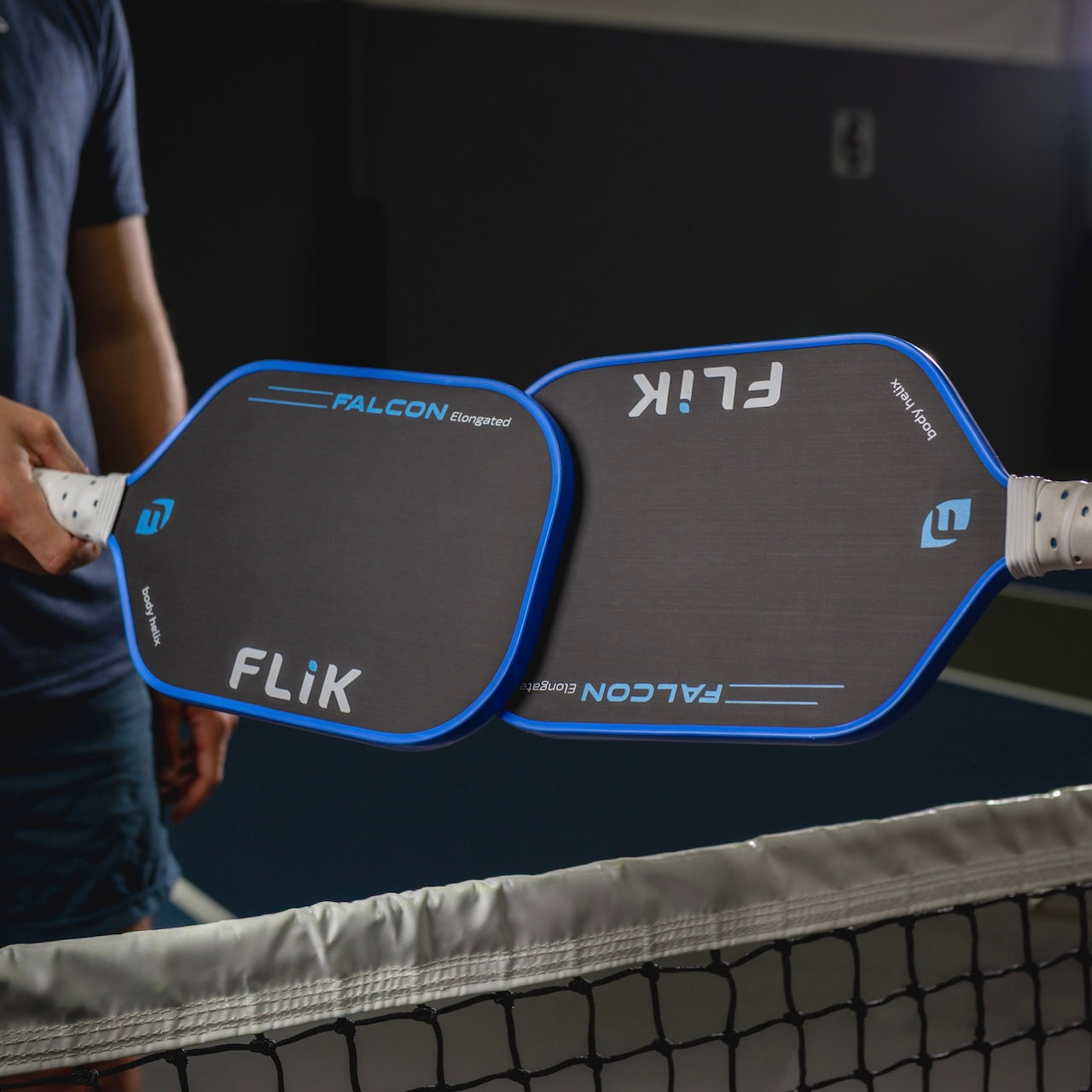 Two people hold blue and black body helix FLiK Falcon Elongated USAP Approved pickleball paddles over a net, paddle faces touching. Indoors, with a blurred background, this highlights the precision and control these paddles provide.