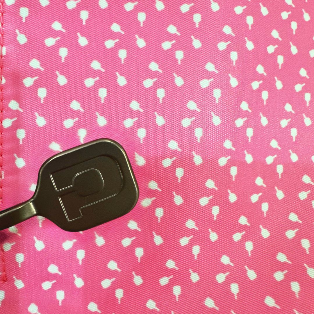 A black key with a rounded rectangular head rests on the pink, teardrop-patterned lining of the Lulu Pickleball Tote from Playly Store, with a stitched seam visible on the left in bright lighting.