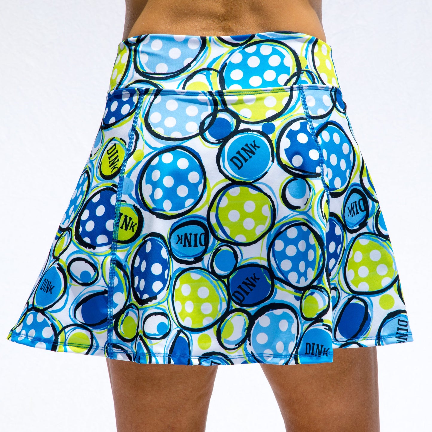 The Pickleball Bella PB Aline Skort Dink 1 is a colorful, flared skirt with blue, green, and white polka-dot circles and "DINK" in bold letters, shown on a model against a plain light background.