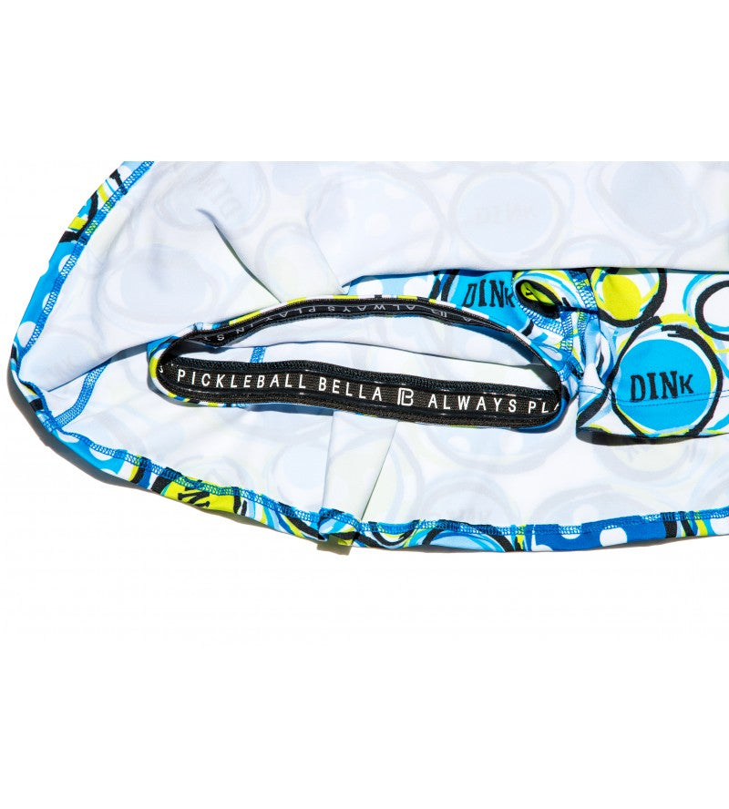 Close-up of the PB Drop Pleat Skort Dink 1 by Pickleball Bella, a flattering white pickleball skirt with blue and green patterns, a black inner waistband reading “PICKLEBALL BELLA ALWAYS PLAY,” and the word "DINK" on one circle.