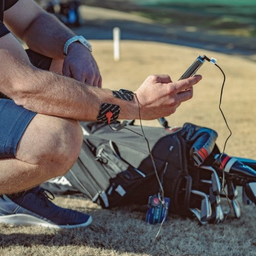 A person crouches on grass near golf bags, using the iReliev SPORT Muscle Stimulator with Explosive Strength Mode—phone in hand, wires connect to patches on their forearm—suggesting use at a golf course.