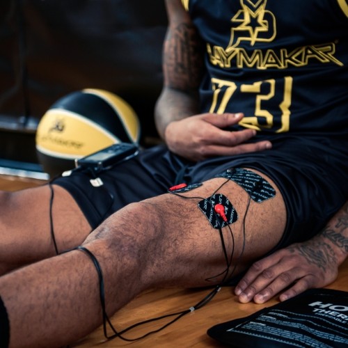 A basketball player in a black and yellow jersey uses the iReliev SPORT Muscle Stimulator with Explosive Strength Mode, with electrodes on their exposed knee, for therapy or recovery. A basketball rests nearby on the wooden floor.