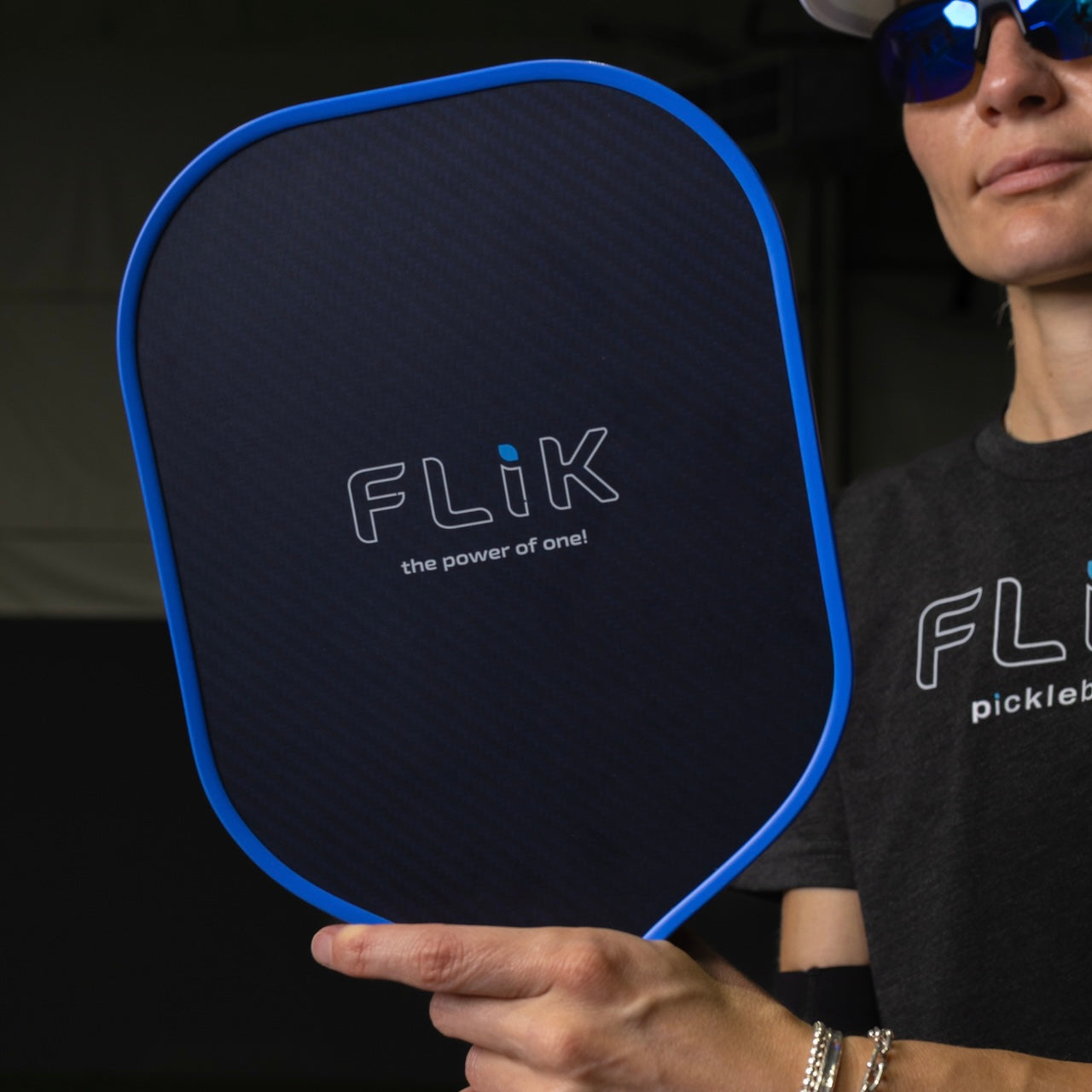 A person in sunglasses holds the body helix FLIK Gen 3 Pickleball Paddle - USAP Approved with a blue edge, wearing a gray FLiK shirt and bracelet. Only part of their face is visible against a dark background.