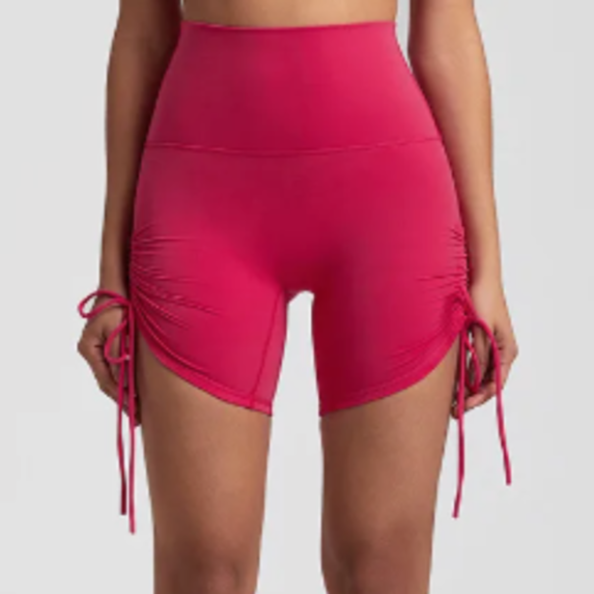 A person models DYM Athletics AIRE Ultra-Soft Ruched Biker Shorts in bright pink, featuring a high waist, adjustable inseam, ruched sides, and anti camel toe design. Image is cropped from lower chest to mid-thigh with arms relaxed.